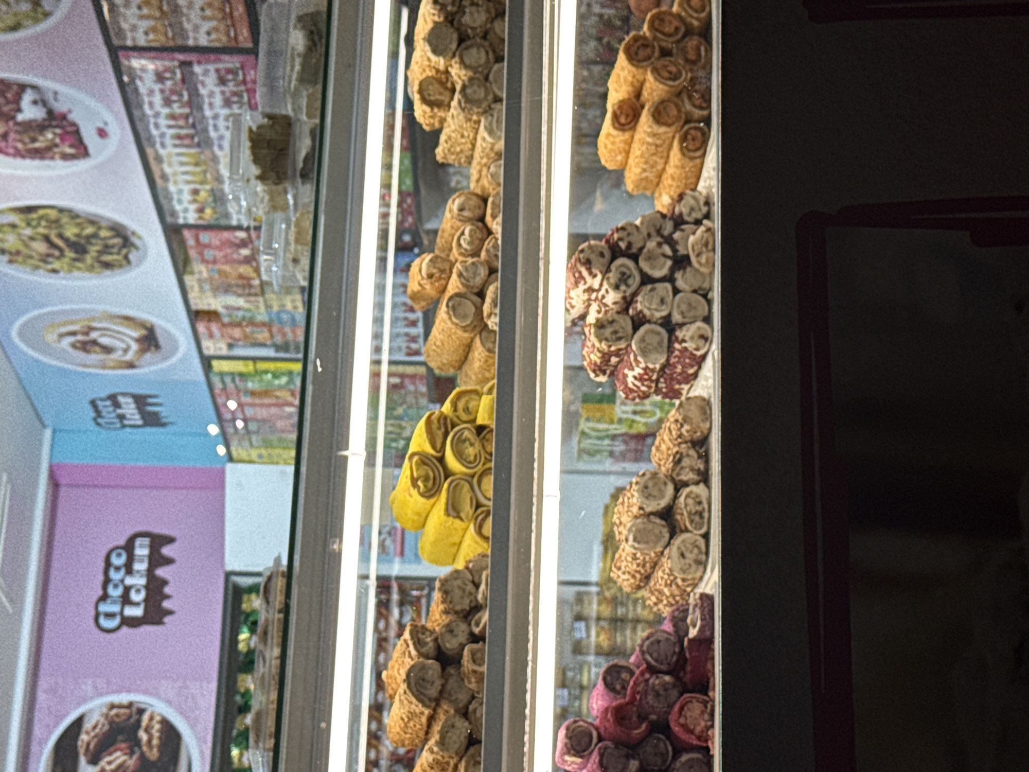 The image shows a display case filled with Turkish delight and other sweets. The sweets are arranged in neat rows and piles, with some of them covered in nuts and others in powdered sugar. The display case is well-lit, and the sweets are colorful and appealing. In the background, there are shelves stocked with packaged sweets and candies. Above the display, a wall features signage that reads "Choco Lokum" and "Lokum," along with circular images of various sweets. The overall impression is of a sweet shop or a confectionary store. This establishment is likely located in Ilidža, Bosnia and Herzegovina, a popular tourist destination known for its natural beauty and historical sites, and where such shops selling traditional sweets are common.