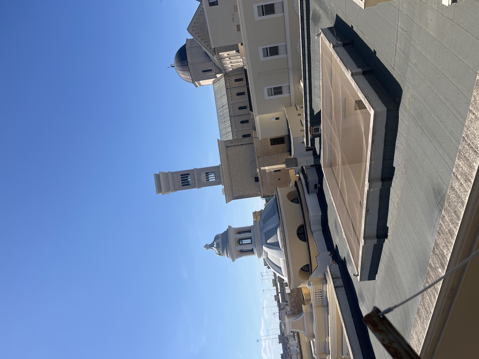 The image is taken from a rooftop in Bisceglie, Italy, at coordinates 41.242, 16.499, looking out at the surrounding cityscape. It is a sunny day with a clear blue sky. The rooftop is made of grey tiles and features a large, rectangular skylight covered in a brown mesh netting. In the background, there are three prominent buildings: a church with a dome, a bell tower with a clock face, and another building with a flat roof and visible windows. The rooflines of other buildings are visible in the distance. The image appears to be taken from a residential building with a yellow facade. The roof is partially obscured by the foreground. There are no people in the scene, and the environment is peaceful.