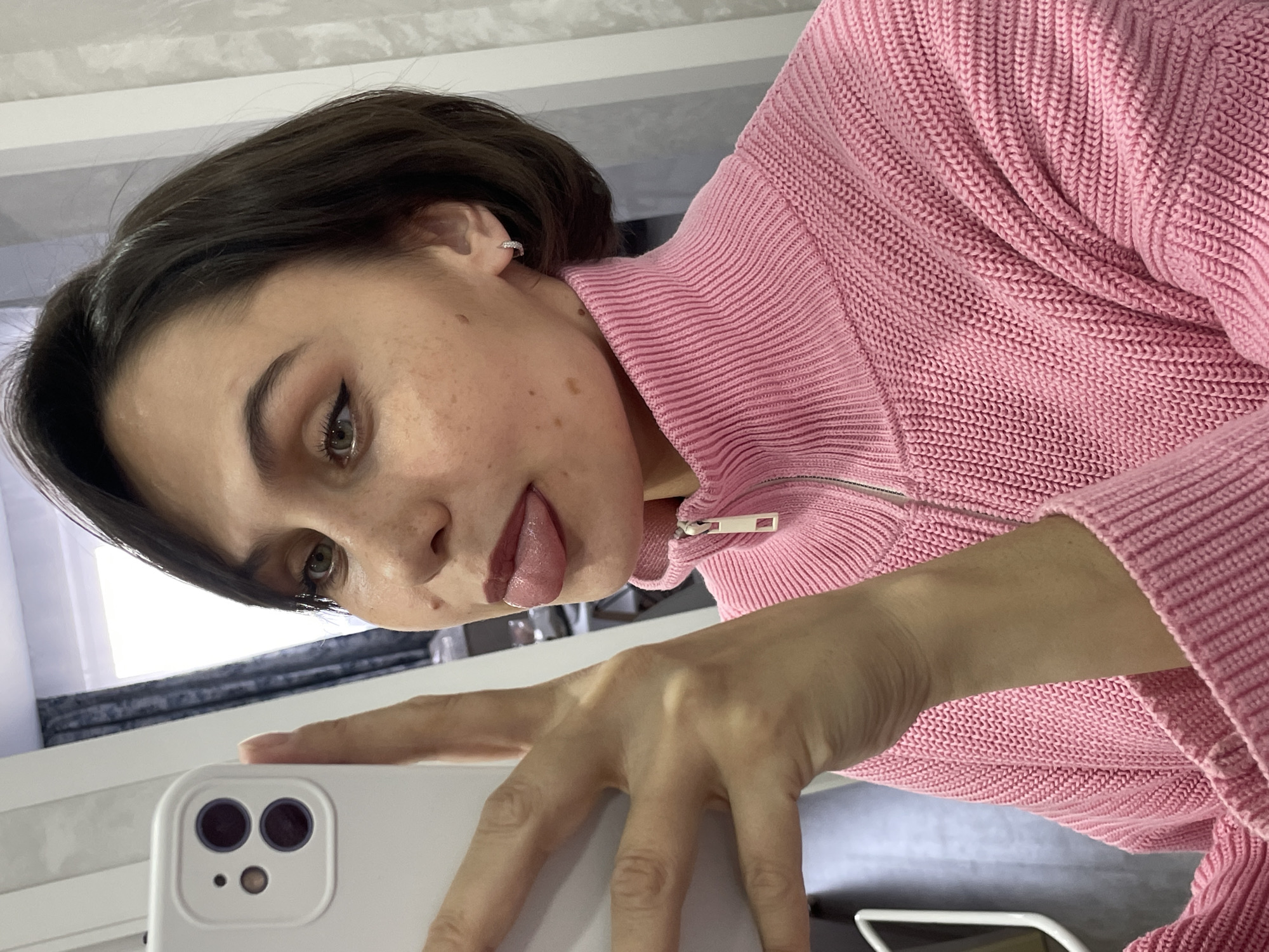 A young woman with short brown hair and fair skin is taking a selfie with a white smartphone. She is wearing a pink knitted sweater with a zipper closure. The woman is sticking her tongue out. She is wearing a small earring in her left ear. The location of the photograph is unknown. The city of Izhevsk, Russia is not ascertainable.