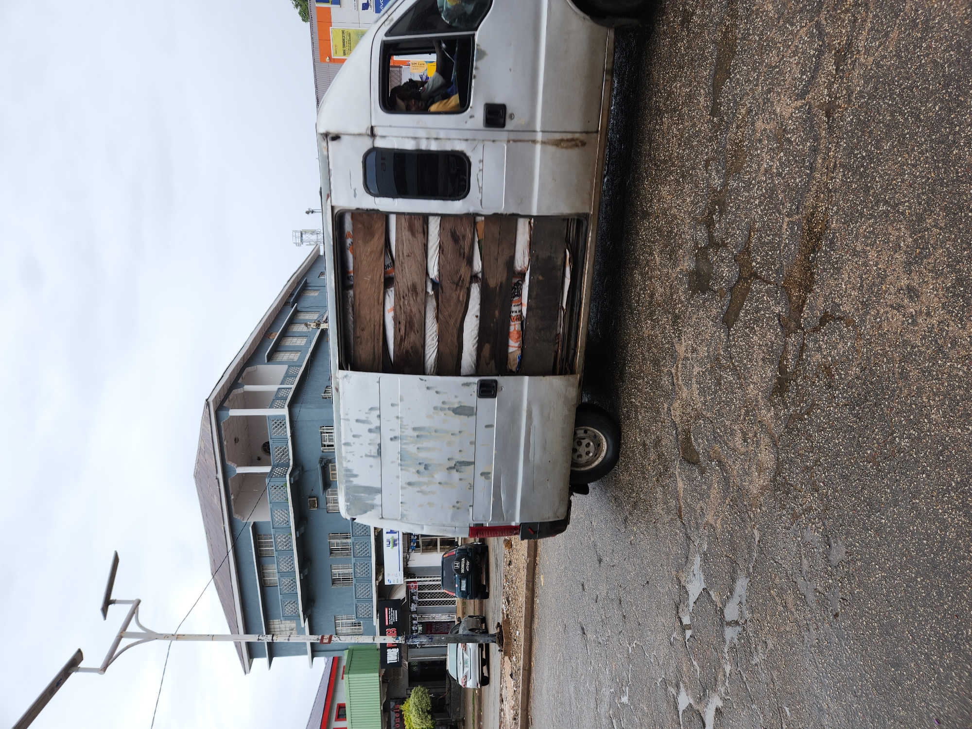 A white van with its side door open is parked on a wet asphalt street in an urban area. The van is heavily loaded with wooden planks and packages. The driver is visible behind the steering wheel. Behind the van, there is a four-story blue building with white trim and multiple windows. Parked cars are visible along the street, and a green and red structure with a sign is partially visible. A street light stands near the van. The location is the city of Unknown, Unknown.