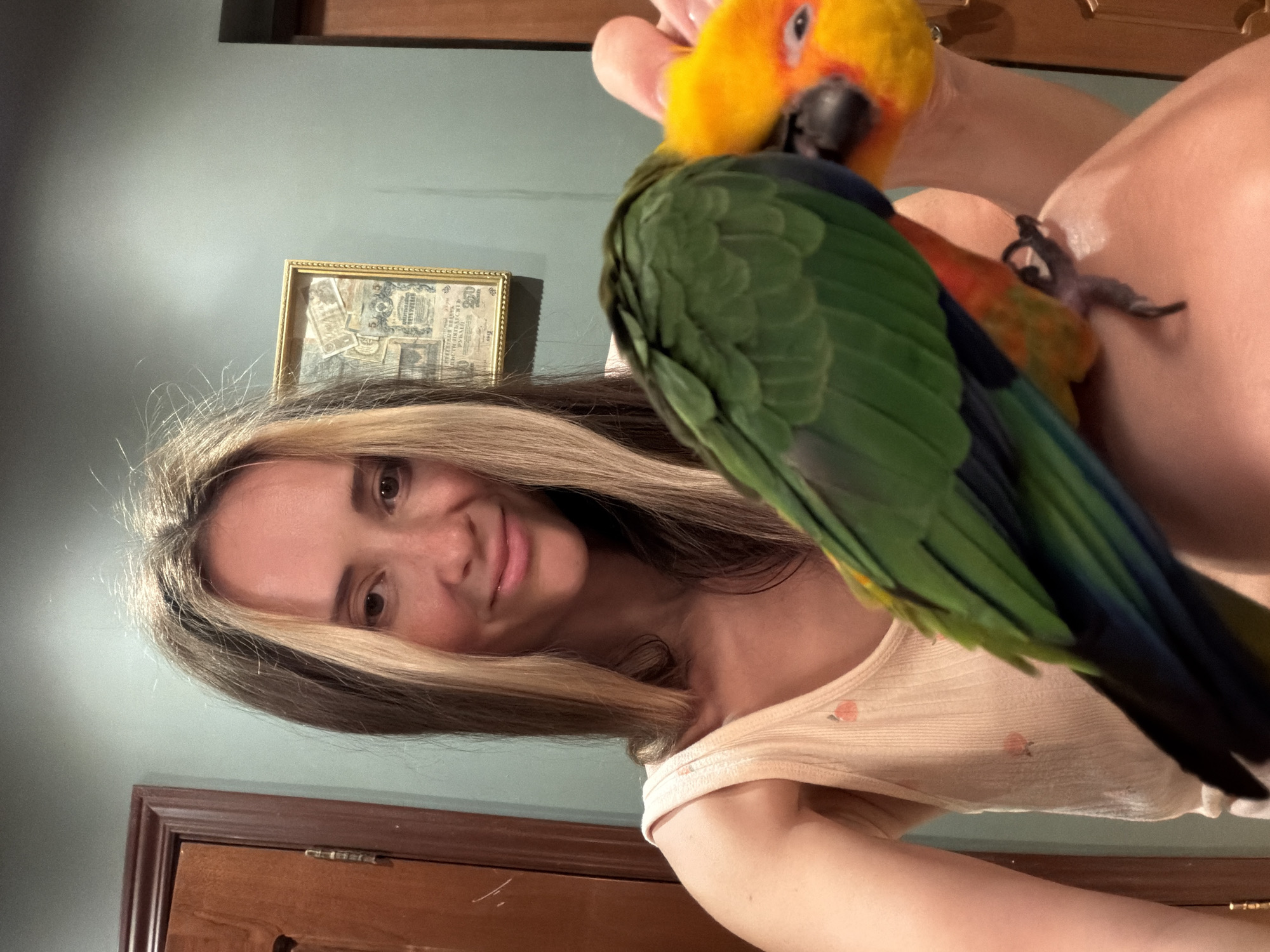 In an indoor setting with teal-colored walls and wooden trim, a woman with long, blonde-streaked brown hair and a light-colored tank top smiles at the camera. She is holding a green and yellow parrot. The parrot is perched on her shoulder and arm, with its head tilted down towards her. A framed picture containing old paper currency hangs on the wall behind the woman. The scene does not provide any information to suggest a location in the city of Khimki, Russia.