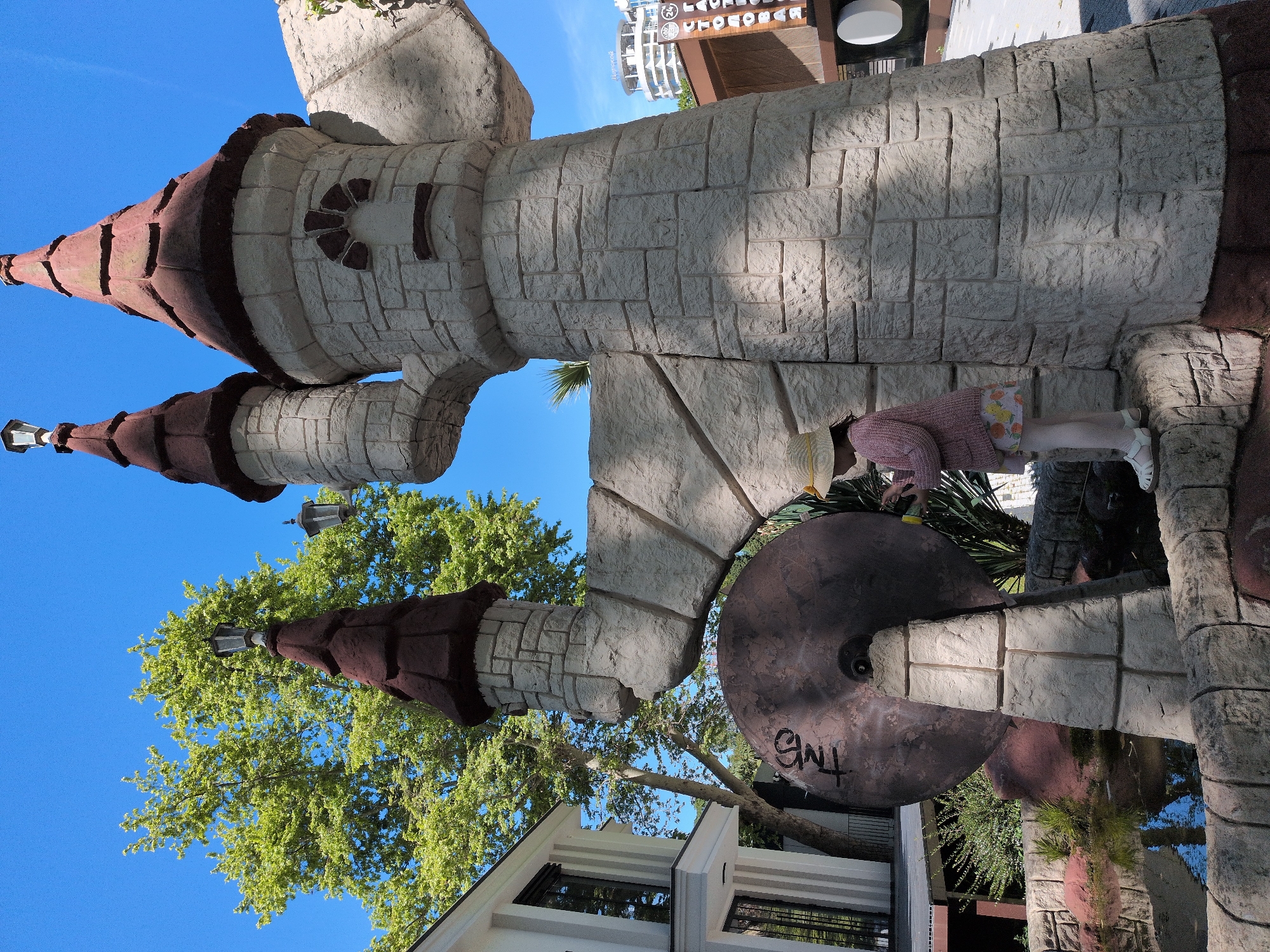The image, captured in Sochi, Russia, depicts an outdoor scene centered around a large, light-colored, stone-textured fantasy castle structure. The castle features multiple turrets with brown, pointed roofs, one of which includes a lamppost. The walls are designed to resemble irregular stone blocks.
A young child or small person is visible on the right, seen from behind, wearing a pink long-sleeved top, patterned shorts, and white sandals, with a straw hat. They are bending over, appearing to interact with the base of the castle structure.
Near the base of the castle on the left, there is a large, dark, circular object, possibly a decorative disc or mill wheel, which has the white graffiti "GNT" written on it.
The scene is set under a clear, bright blue sky, indicating sunny weather. Lush green trees, including deciduous trees and palm fronds, surround the castle, with some appearing in the background. Modern buildings are visible in the distance, one of which prominently displays the text "LAURA" along with other partially obscured letters. Strong shadows cast by the structures suggest a bright day, likely midday or early afternoon. The overall environment suggests a recreational area, such as a resort or park.