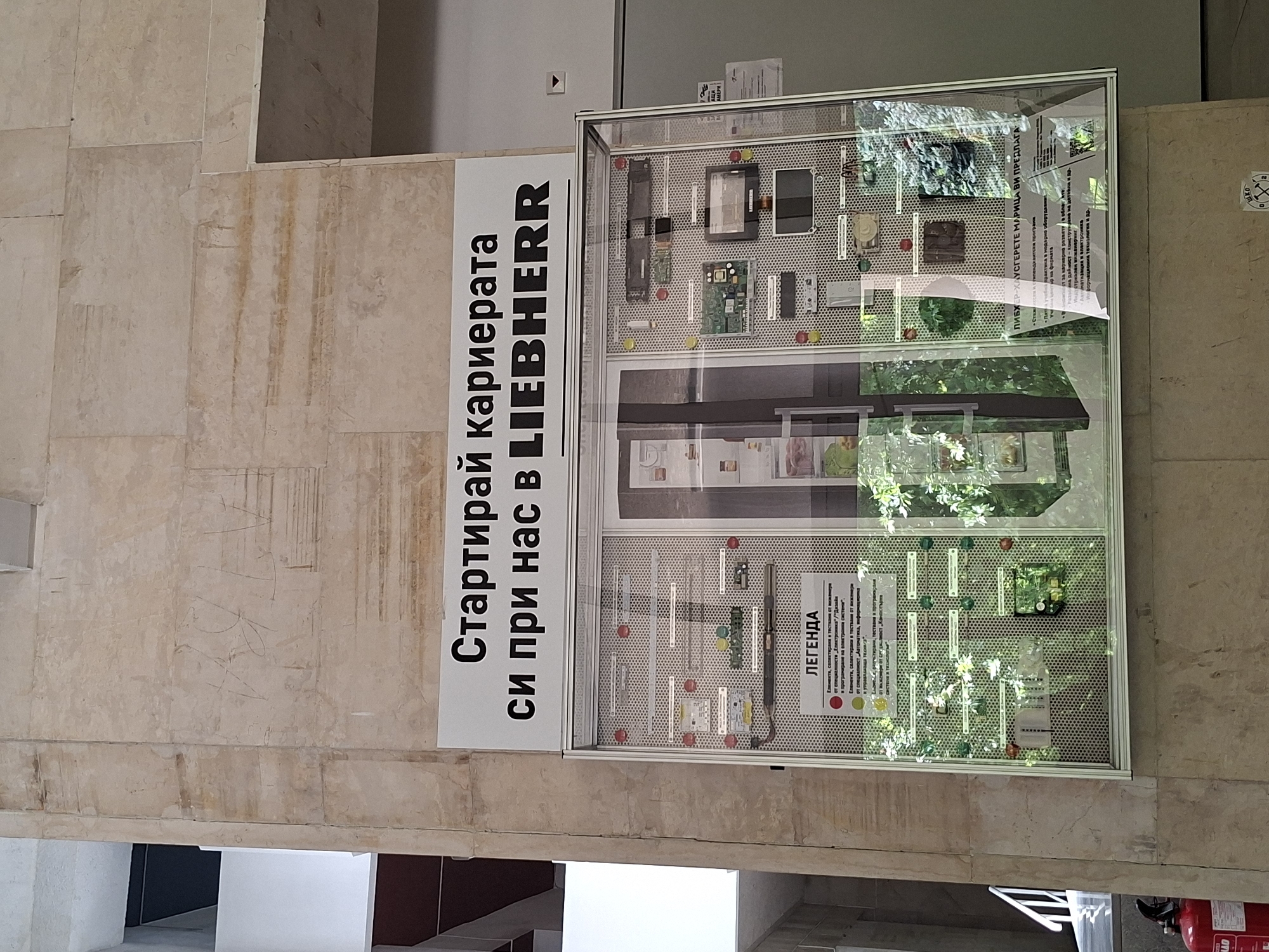 The image displays an indoor scene featuring a promotional display for Liebherr, likely within a company building or an exhibition area. On the left, a large white sign is affixed to a textured, light brown tiled wall. The sign, oriented vertically, prominently reads in Bulgarian: "Стартирай кариерата си при нас в LIEBHERR" (Start your career with us at LIEBHERR).
Adjacent to the sign on the right is a glass display case, containing various electronic and mechanical components mounted on a perforated metal panel. These items appear to be parts, sensors, circuit boards, and other technological elements relevant to engineering or manufacturing. Reflections on the glass case show greenery, possibly from plants or outdoor elements, and internal structural reflections.
Inside the display case, text is visible, partially obscured by reflections. The top section, under the heading "ЛИБХЕР-ХАУСГЕРЕТЕ МАРИЦА ВИ ПРЕДЛАГА" (LIEBHERR-HAUSGERÄTE MARITZA OFFERS YOU), lists career development opportunities in fields such as engineering, automation, electronics, and IT. The bottom section presents a "ЛЕГЕНДА" (LEGEND) using colored markers (red, yellow, green) to categorize the displayed components. Red markers indicate elements selected and tested by engineers in "Electronics" and "Design." Yellow markers denote elements from "Automation, Information Systems, and Technologies." Green markers identify elements from "Mechatronics" and "Production Process Management."
The setting is an interior space with warm-toned, large rectangular tiles covering the walls, suggesting a professional or corporate environment in Plovdiv, Bulgaria.