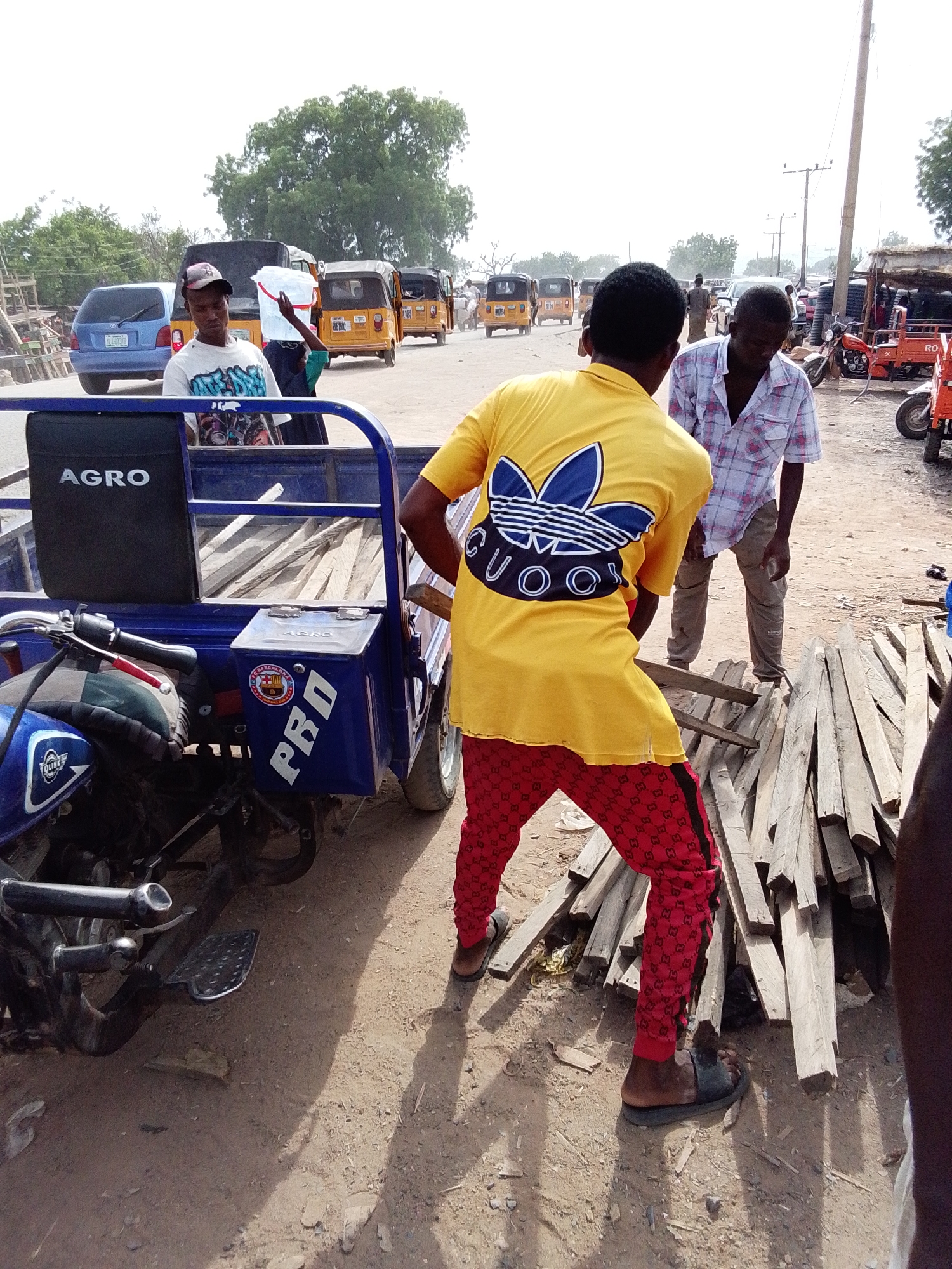 The image depicts a street scene in Jimeta, Nigeria. Two men are standing near a pile of wooden planks, next to a blue tricycle that is parked on a dirt road. The man closest to the camera is wearing a yellow t-shirt with a blue graphic and red patterned pants with black stripes. The other man is wearing a light-colored plaid shirt and brown pants. In the background, there are several yellow auto-rickshaws, a blue car, and other people. Power lines are visible above. The environment appears to be a busy commercial area.