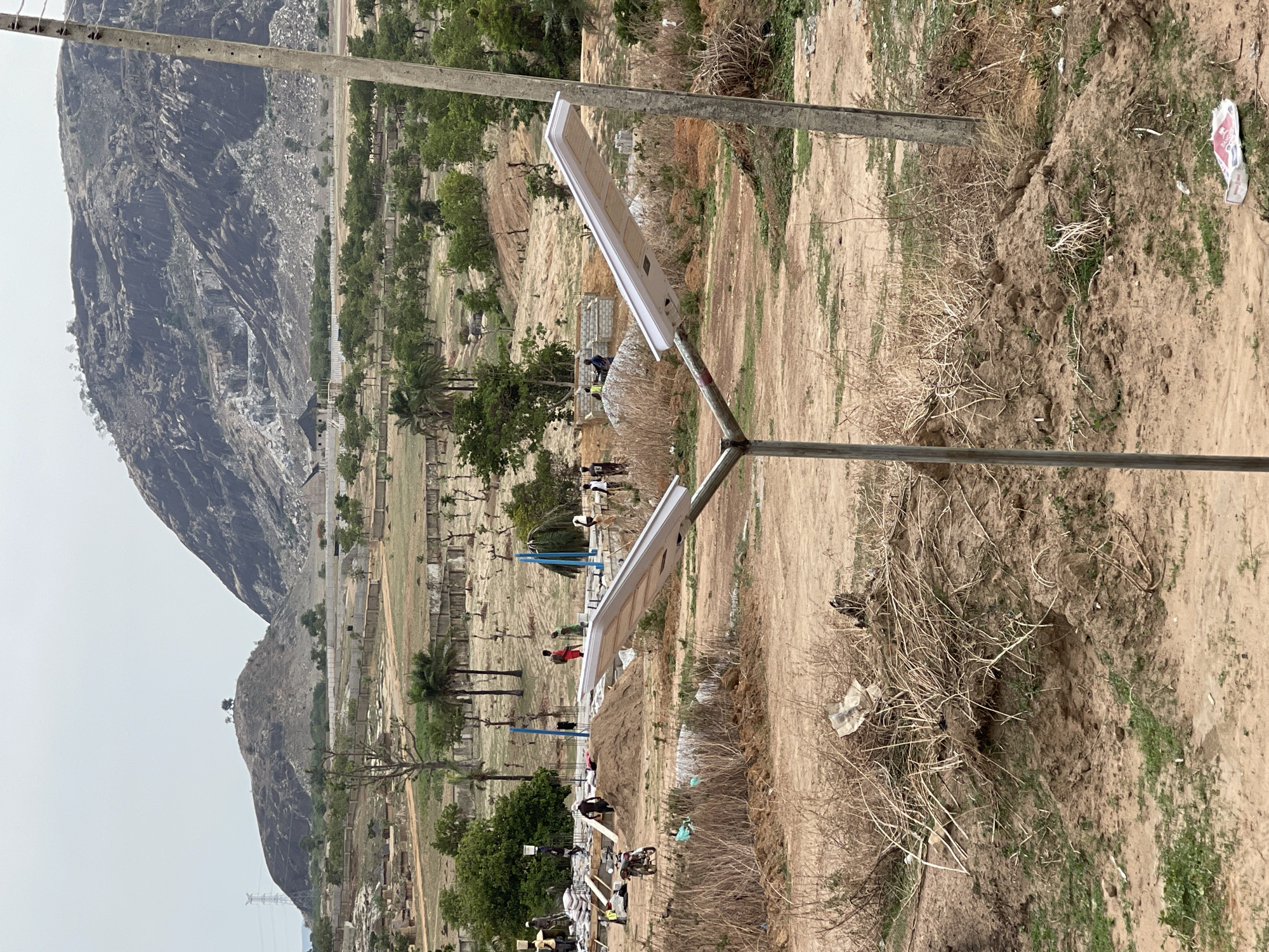 The image depicts a scene in Angwan Sayawa, Nigeria, featuring a landscape with a prominent rocky mountain in the background. The foreground shows a dusty, dirt-filled environment with sparse vegetation, including dry bushes and some patches of green. A utility pole-like structure with two rectangular, white, solar-powered lights extends into the scene. In the middle ground, there are several people visible, some appearing to be involved in construction or building activities, as evidenced by what appears to be lumber and construction materials nearby. A playground with blue metal structures is also visible in the distance. There is also a motorcycle at the construction site.