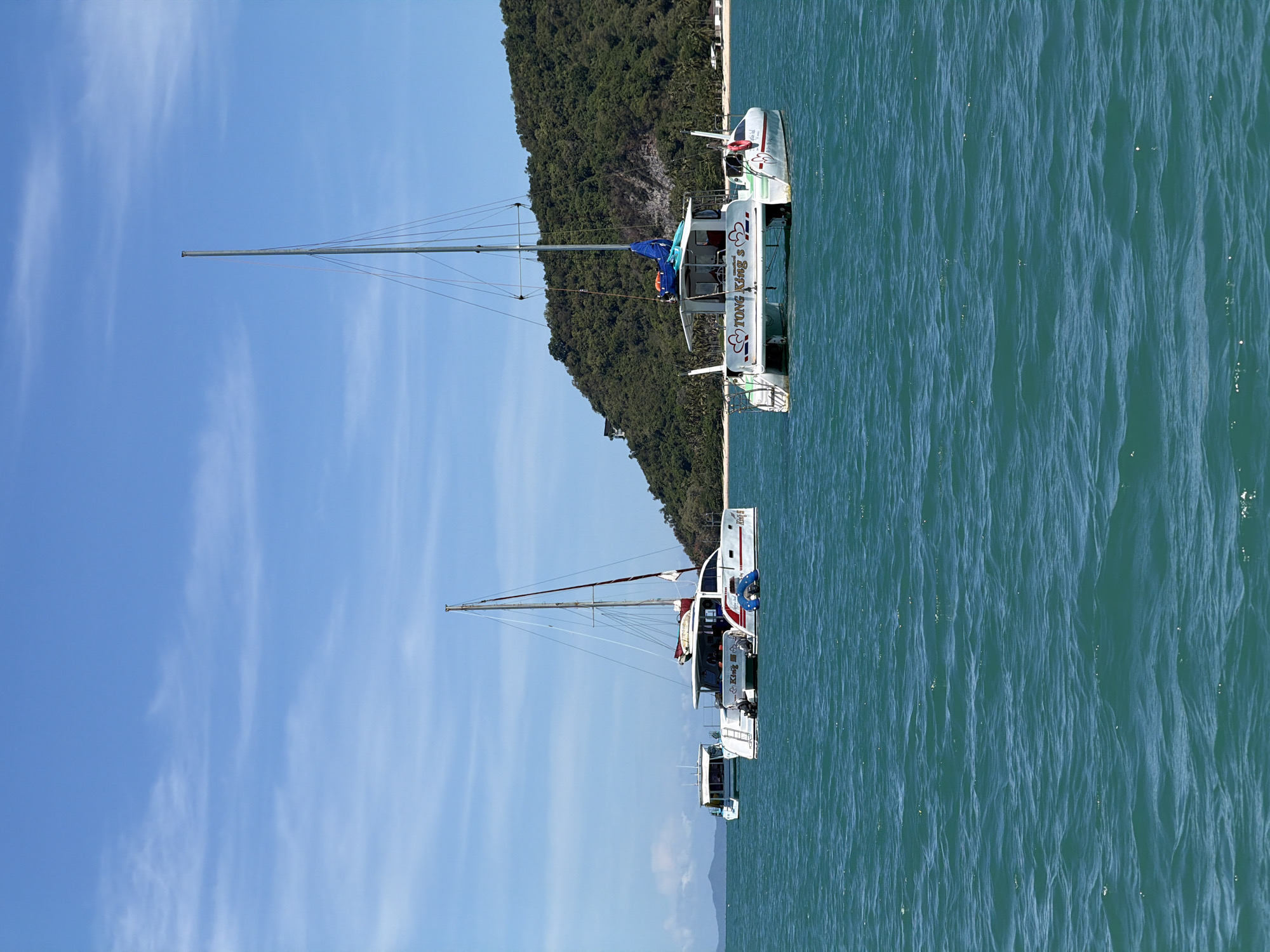 A vertical shot shows two white catamarans anchored in the turquoise waters of Baan Thong Krut, Thailand. The catamaran in the foreground has a blue sail and the words "TONG KING S" written in red on its side. The other catamaran, positioned slightly behind and to the left, also displays white and red markings. In the background, a lush green, tree-covered island rises from the sea, with hints of buildings visible on its slopes and along the shoreline. The sky is a clear blue with a few wispy white clouds. The water is calm with gentle ripples.