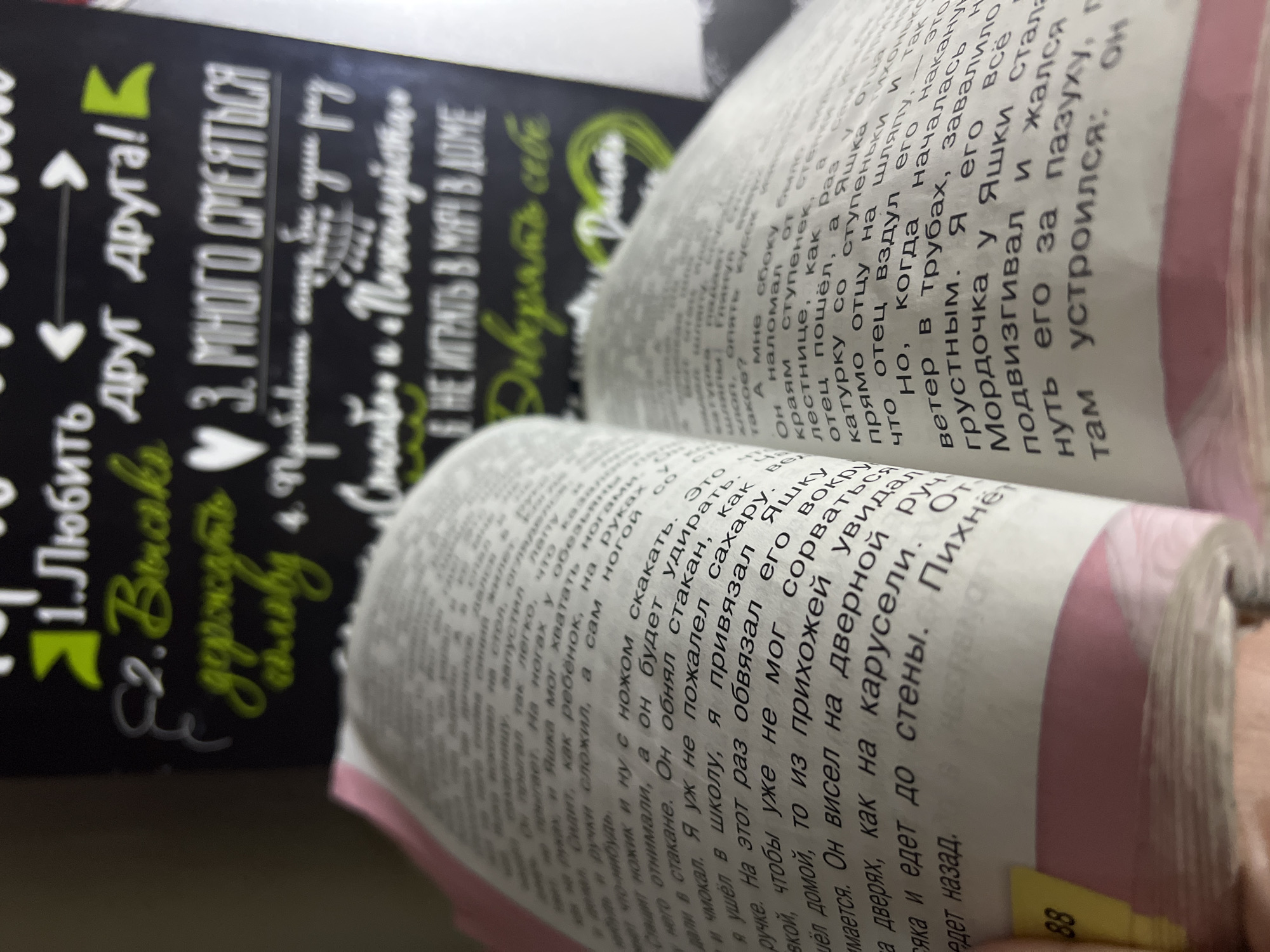 The image depicts an open book with Russian text, positioned in front of a chalkboard-style sign also featuring Russian text and graphics.
The book's pages are filled with text, partially blurred, but legible enough to discern a narrative, possibly a story or novel. The binding is visible along the spine. A yellow sticky note is visible on the right page.
Behind the book is a black chalkboard sign with white and green text and graphics. The text appears to be a list of suggestions or rules.
The environment seems to be indoors, likely a home or office setting, given the objects present.
The location is assumed to be Noviy, Russia, based on the language used in the text.