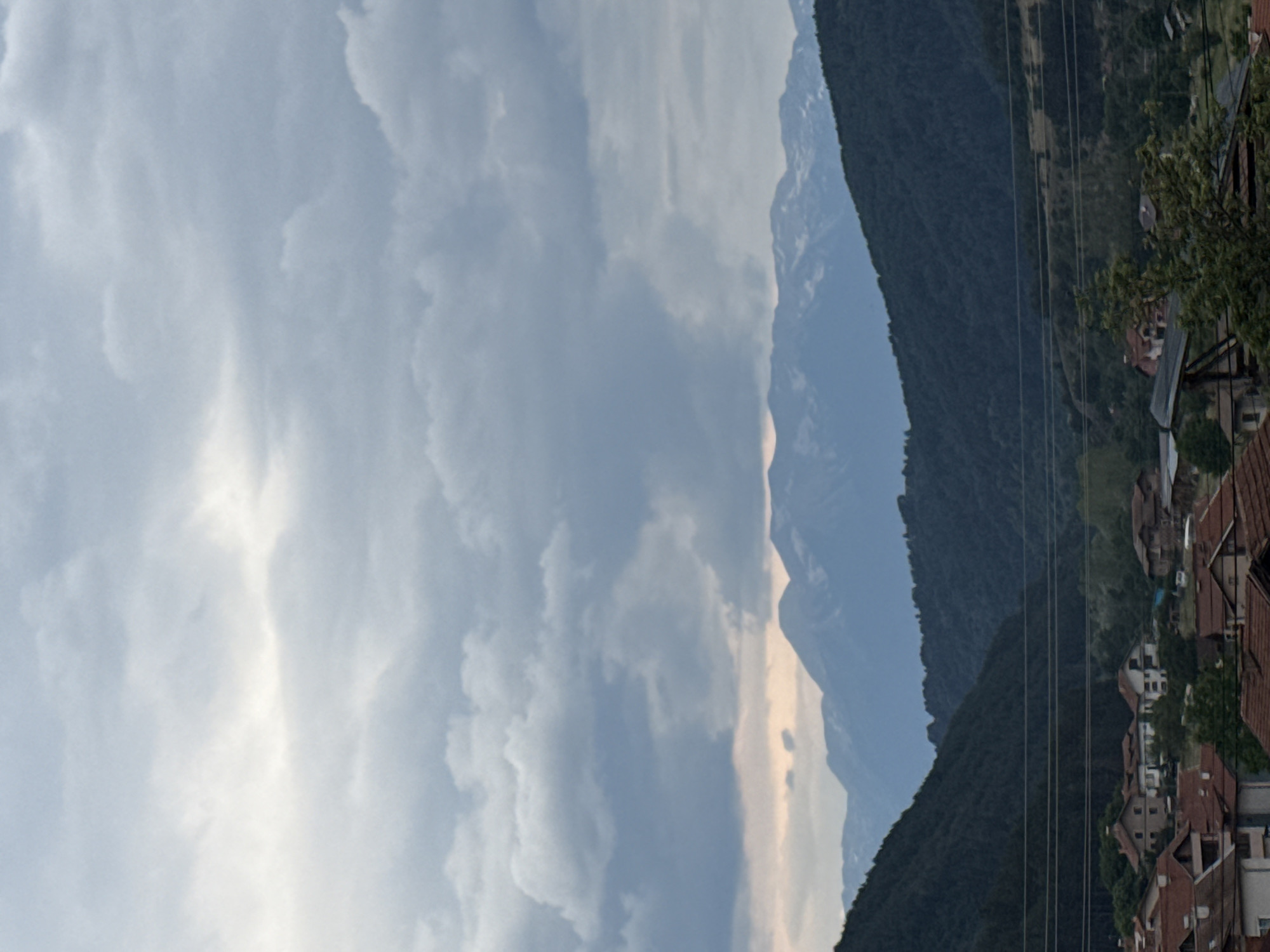 The image depicts a landscape scene with a village in the foreground, nestled at the foot of rolling hills and majestic mountains. The sky is overcast with dramatic clouds, suggesting either dawn or dusk, with a faint glow of sunlight breaking through in the upper left quadrant.
In the foreground, the village of Yurukovo, Bulgaria, is visible, characterized by a cluster of houses with red-tiled roofs. The architecture appears traditional, with some buildings showing signs of age and others looking more recently constructed. Trees are interspersed among the houses, and power lines are strung across the midground, indicating the presence of infrastructure.
Beyond the village, the landscape transitions into dark, densely forested hills that rise steeply. These hills create a dramatic backdrop, framing the view of the distant mountains. The mountains themselves are formidable, with their peaks dusted with snow, indicating a higher altitude and a colder climate. The overall impression is one of a remote, mountainous region with a quiet, rural settlement.
There are no people visible in the image, nor any explicit actions or interactions taking place. The scene is static and focuses on the natural beauty and the harmonious coexistence of the village with its surrounding environment. The lighting is soft and diffused, characteristic of an overcast sky, contributing to a somewhat moody and atmospheric quality of the image.