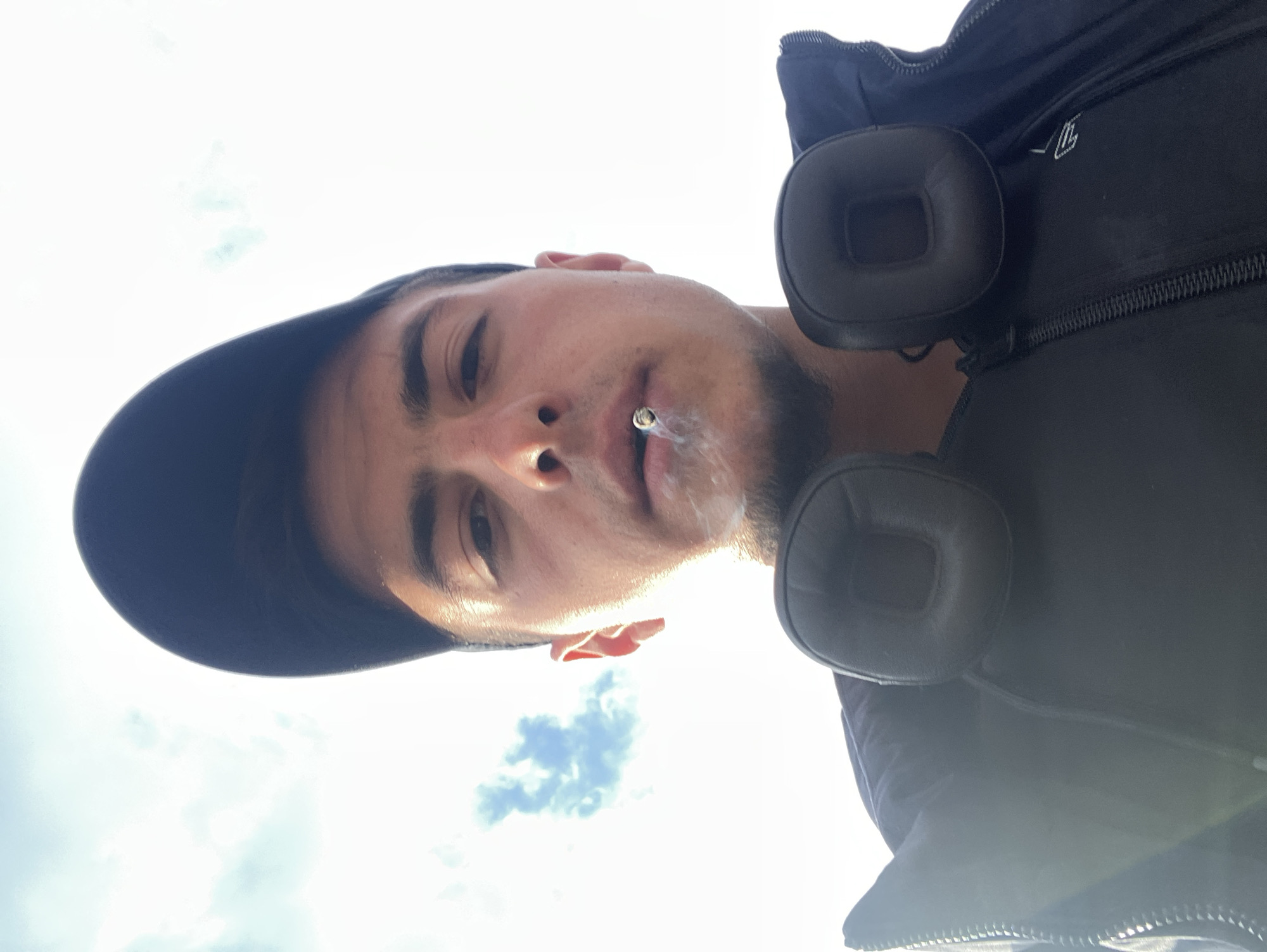 A selfie-style shot is taken from a low angle, looking up at a person with fair skin, dark hair, and a slight beard. The person is wearing a dark-colored baseball cap and has what appears to be a cigarette or similar item in their mouth, emitting smoke. The background consists of a bright sky with some clouds visible. The person is also wearing a dark jacket or vest, with a pair of over-ear headphones resting on their chest. There is no information about the city of Krastevich, Bulgaria present in the image.