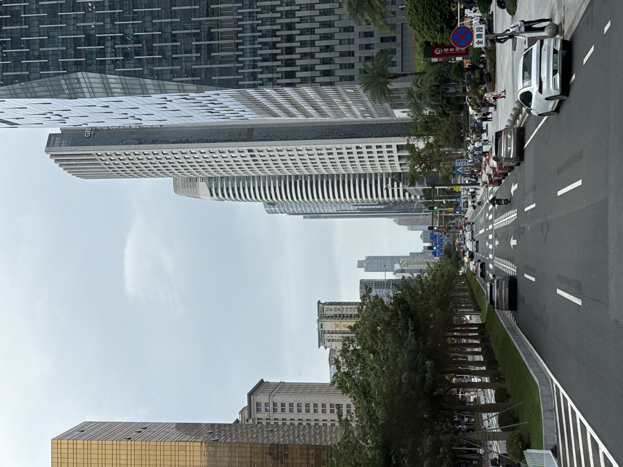 The image depicts a street in the Tianhe District of Guangzhou, China. The street runs between tall modern buildings, some with distinctive architectural features like gridded facades and curved elements. The road is paved and divided into lanes with white markings and arrows indicating direction. Cars and other vehicles are visible on the road, with a few parked on the side. On the sidewalk, people are walking. A row of trees lines the side of the street, separating the sidewalk from the road. A few traffic signs are visible, one with Chinese characters. The sky is overcast.
