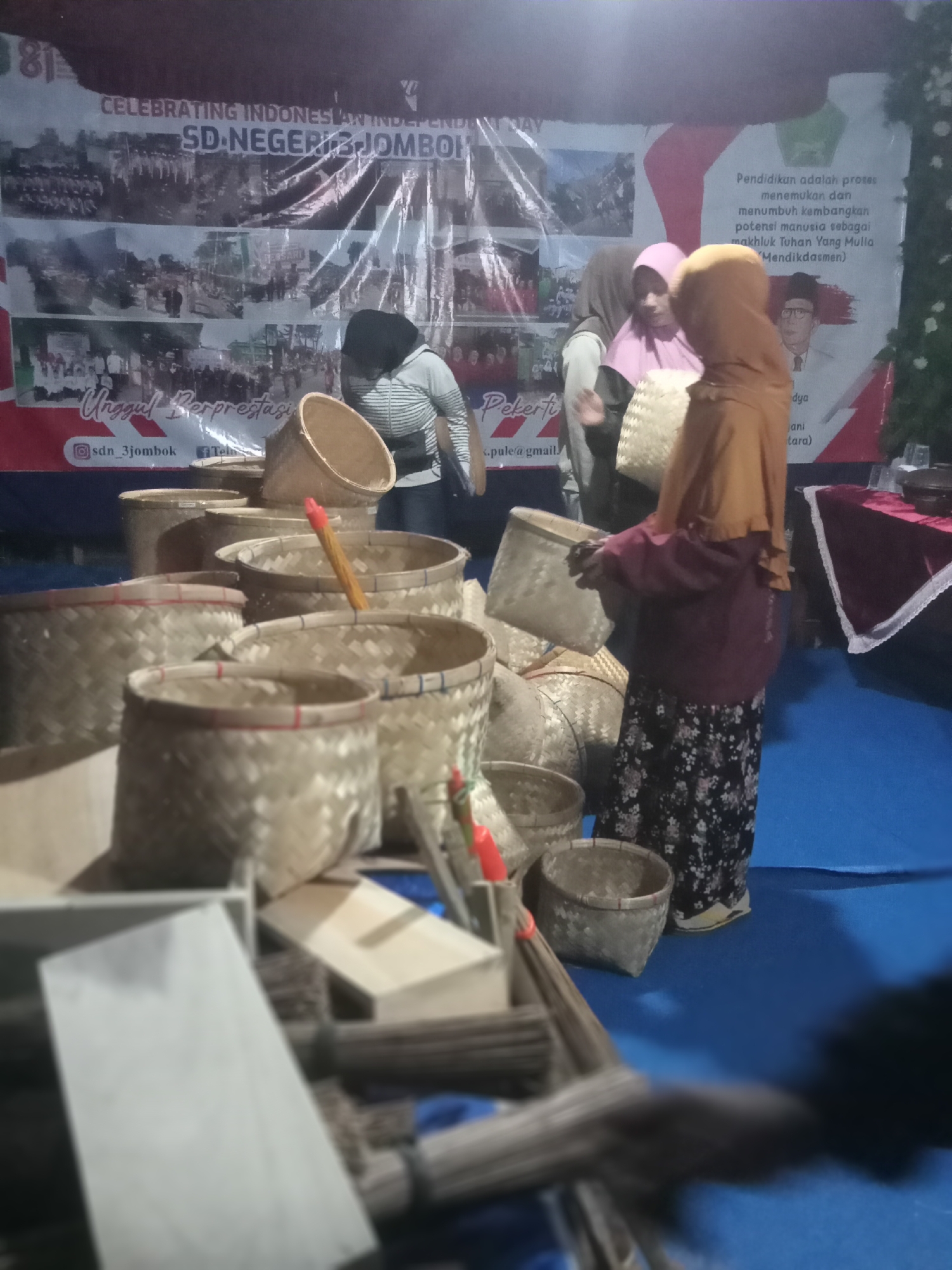 The image depicts an indoor scene, likely a market or exhibition, with a prominent banner in the background reading "CELEBRATING INDONESIAN INDEPENDENCE SD NEGERI 3 JOMBOK". The banner features multiple photographic panels. In the foreground, there are numerous woven bamboo baskets of various sizes, arranged in stacks and clusters. A few individuals are visible, most notably a woman in a tan headscarf and a maroon long-sleeved shirt over a floral patterned skirt, holding one of the baskets. Another woman, partially obscured, is wearing a striped shirt and a dark headscarf. The environment appears to be a stall with a blue floor covering. The overall impression is of a display of local crafts or products.