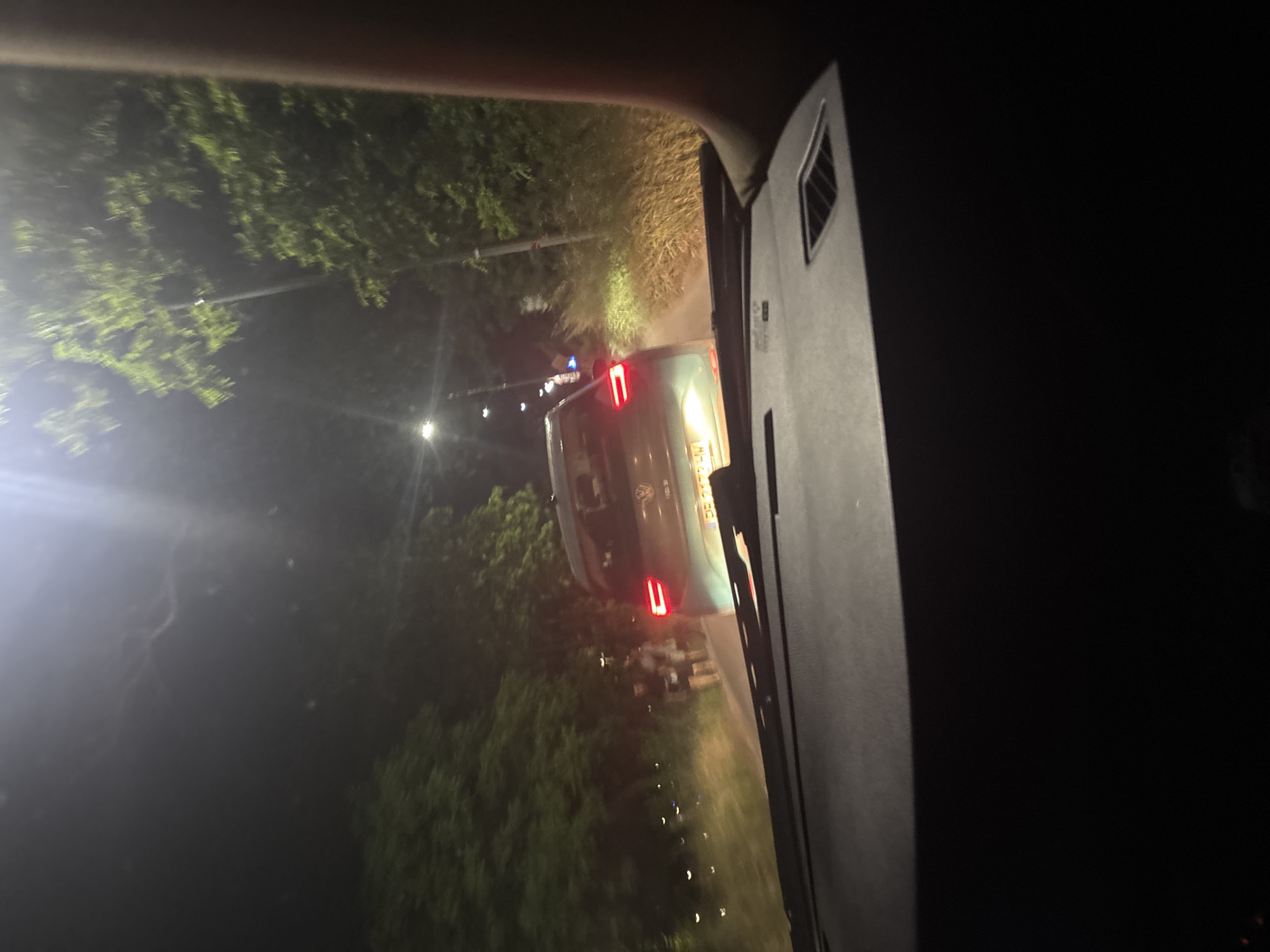 The image appears to be taken from inside a vehicle, possibly a car, at night. Through the windshield, one can see the rear of another vehicle, a van, with its tail lights illuminated. The van seems to be traveling on a road lined with trees on both sides. Streetlights are visible along the road, casting a glow on the scene. The location appears to be somewhere in Plovdiv, Bulgaria. The interior of the vehicle from which the photo was taken is dark, with a side view mirror visible on the right side of the frame.