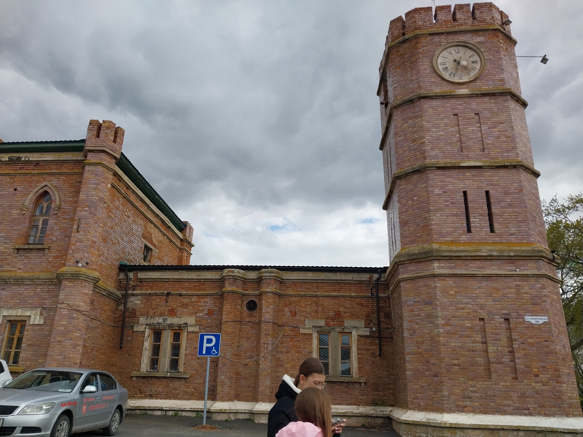 This media file captures a distinctive brick building with a prominent clock tower under an overcast, cloudy sky. The building exhibits a historical architectural style, characterized by reddish-brown brickwork, arched windows with wooden frames, and crenellated parapets on both the clock tower and a section of the main structure. The clock tower features a round clock face with Roman numerals, which appears to indicate a time around 12:45 or 1:00.
In the foreground, two individuals are partially visible, with their backs mostly to the camera. The taller person, wearing glasses and a black jacket, seems to be looking downwards, possibly at a mobile device. A younger person in a pink top stands beside them.
To the left, a silver Skoda sedan is parked. Visible text on the car includes "56-59-26" and "ВЫКУП-1". A blue square sign with a white "P" and a wheelchair symbol designates accessible parking near the building.
On the base of the clock tower, a small white sign clearly displays the address: "ул. НАБЕРЕЖНАЯ" (Naberezhnaya Street) and the number "29". The setting is an urban area in Orenburg, Russia, with the overcast sky contributing to a somewhat subdued atmosphere.