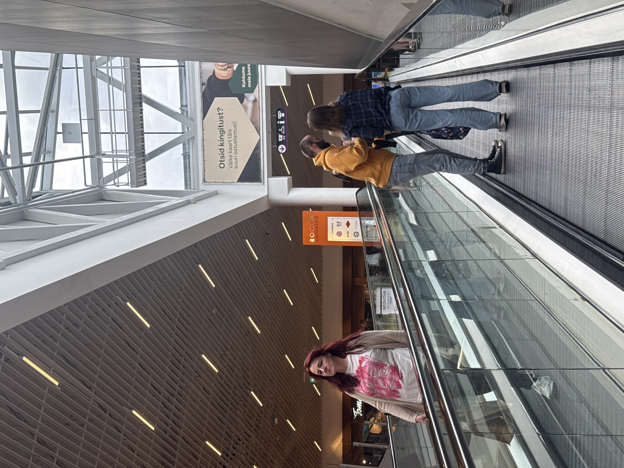 This image captures an indoor scene, likely a shopping mall or airport, with a focus on escalators and the people using them. The architecture features a high ceiling with a slatted metallic structure and overhead lighting. In the foreground, two people, a man wearing a yellow hoodie and jeans, and a woman in a plaid shirt and jeans, are standing on an escalator, facing each other in conversation. The woman is holding a patterned bag. To the left, a woman with long red hair, wearing a light-colored cardigan over a t-shirt with a pink graphic, is ascending on another escalator. She is looking towards the left side of the frame. Reflections in the glass barriers of the escalators show other individuals and the surrounding environment. In the background, there are shop facades, including one with an orange sign that reads "ROCCA". A large advertisement banner is visible on a wall, featuring text in Estonian which appears to be a question about finding a gift. The overall atmosphere is that of a busy public space. While the specific location in Tallinn, Estonia cannot be definitively determined from the visual cues alone, the presence of Estonian text on signage suggests a connection to that country.