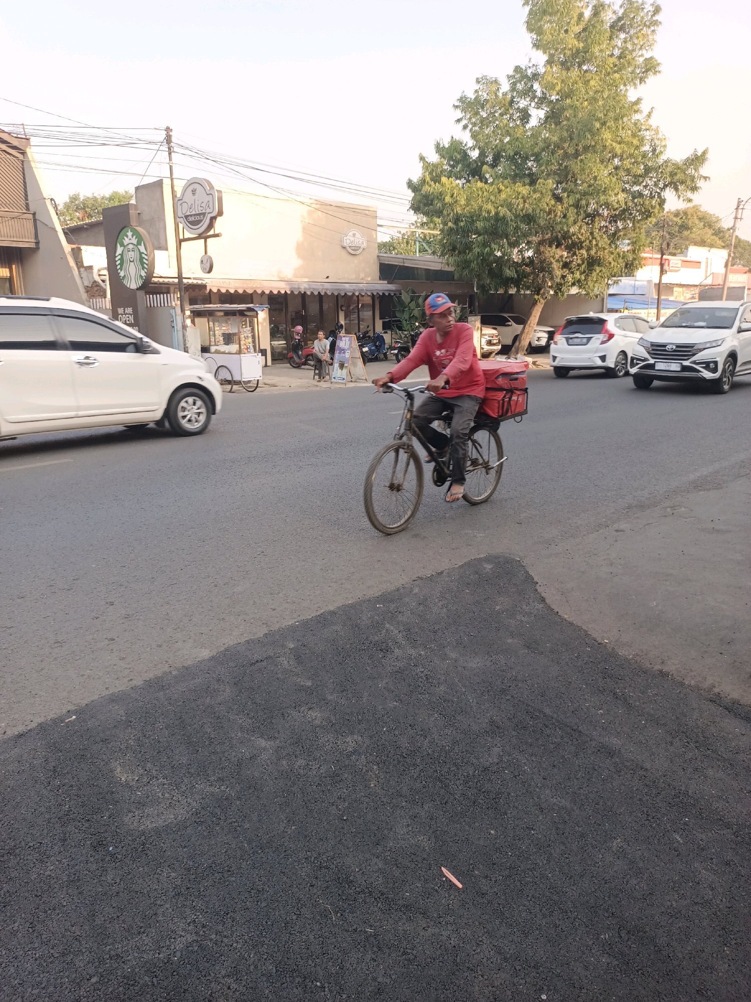 A man is riding a bicycle on a paved road in Purwakarta, Indonesia, in the afternoon. He is wearing a red long-sleeved shirt, dark pants, flip-flops, and a red, blue, and yellow baseball cap. The bicycle has a red insulated delivery box mounted on the rear rack. In the background, there are several white cars, a building with a "Delisa delicious" sign, and a Starbucks sign. There is also a large tree and overhead power lines. The road is partially covered with a fresh layer of dark asphalt.