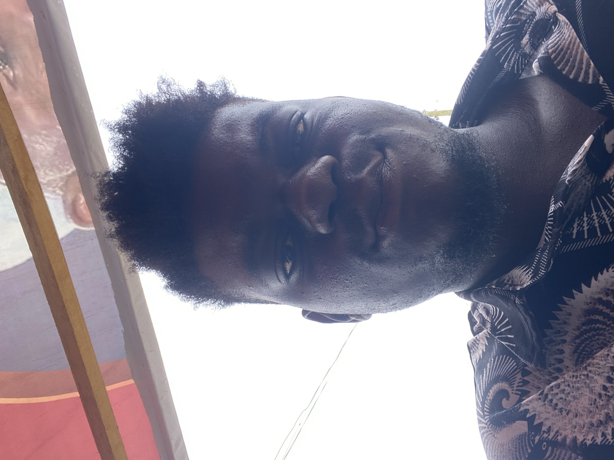 A low-angle close-up shot of a man with dark skin and short, dark, curly hair. He has a slight beard. The background is bright white, suggesting daylight or bright artificial light. The left side of the frame shows part of a structure, including vertical posts with a visible red section. The man wears a patterned shirt. The location is not specified in the city of Unknown, Unknown.