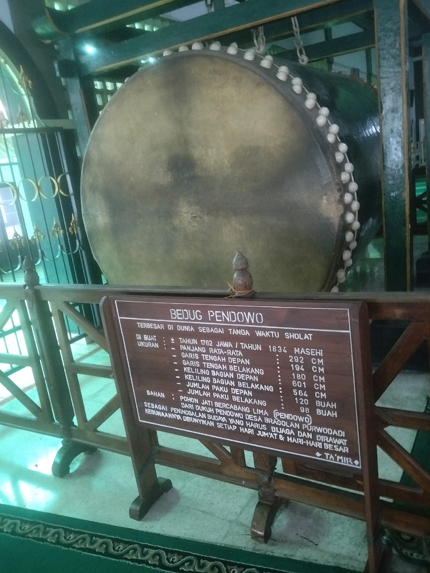 An indoor shot shows a large, weathered drum, possibly a bedug, hung from a dark green metal frame. The drum, with a light brown surface and white beads hanging along its circumference, dominates the upper portion of the frame. In front of the drum, a dark wooden sign with white text details its specifications, including its creation date, dimensions like length and circumference of various parts, and material. The sign mentions "Bedug Pendo" and specifies measurements in centimeters and units of "buah" (fruit, likely referring to the number of decorative elements or fasteners). The environment appears to be a dimly lit, possibly historical building, with some light filtering in from the left. The floor is covered in a green carpet with an ornate pattern. A wooden railing separates the viewer from the drum and sign.