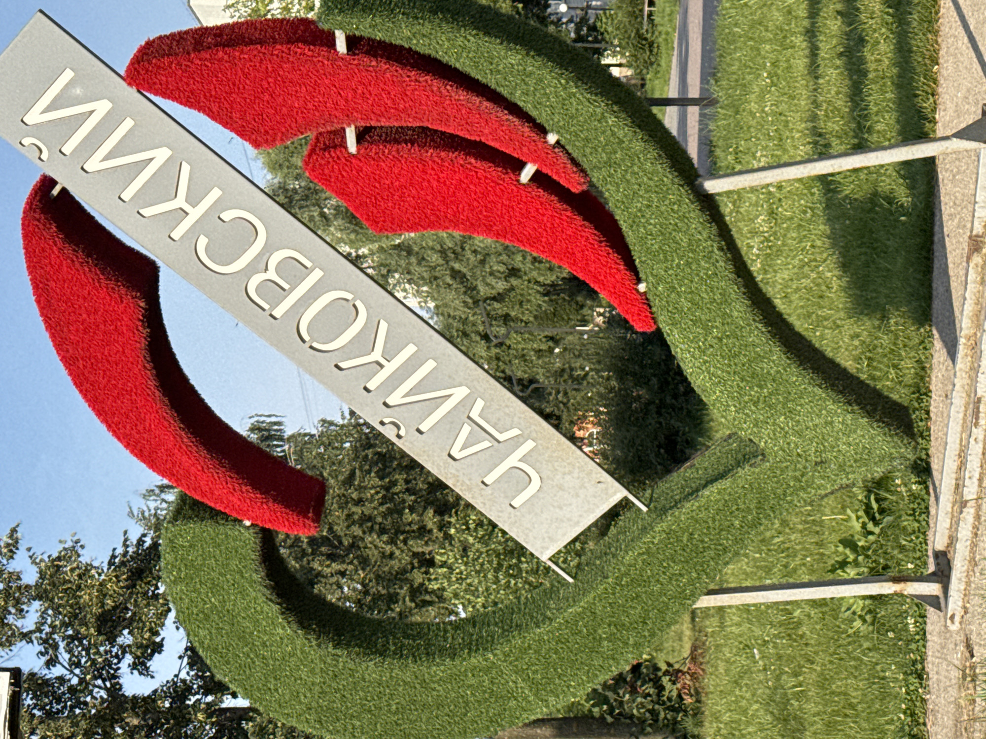 This is a medium shot photograph of a decorative heart-shaped sculpture covered in green artificial turf and red plush fabric, with a metal sign inscribed with Cyrillic lettering. The sculpture is outdoors, in a park or public square, with a background of green trees and a clear blue sky. The sign, which is angled diagonally across the sculpture, reads "ЧАЙКОВСКИЙ". This translates to "Chaikaovsky" or "Tchaikovsky" in English, likely indicating a place or person of significance. The sculpture is supported by metal poles and stands on a paved area, with grass and small white flowers in the foreground. The overall scene suggests a sunny day in an urban or suburban environment. There are no people visible in the image. The sculpture itself appears to be a landmark or public art installation.