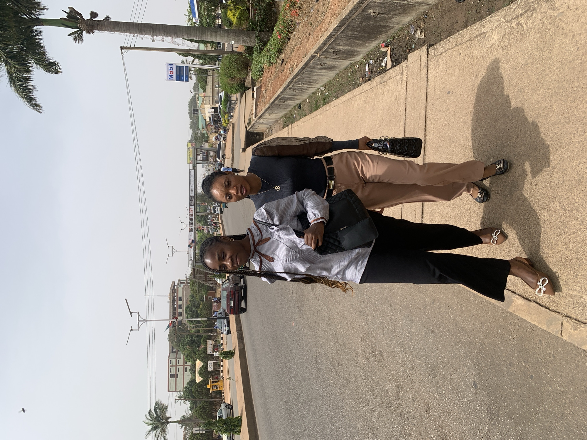 Two women stand on a sidewalk alongside a road in Jos, Nigeria. The woman in the foreground wears a horizontally striped shirt, black pants, and brown flats adorned with white bows. She carries a black shoulder bag. The woman behind her wears a black long-sleeved shirt, tan pants, and black flats. She is holding a shiny black purse. A Mobil gas station sign is visible in the background, among other commercial buildings. The sky is overcast. A palm tree stands to the left of the scene.