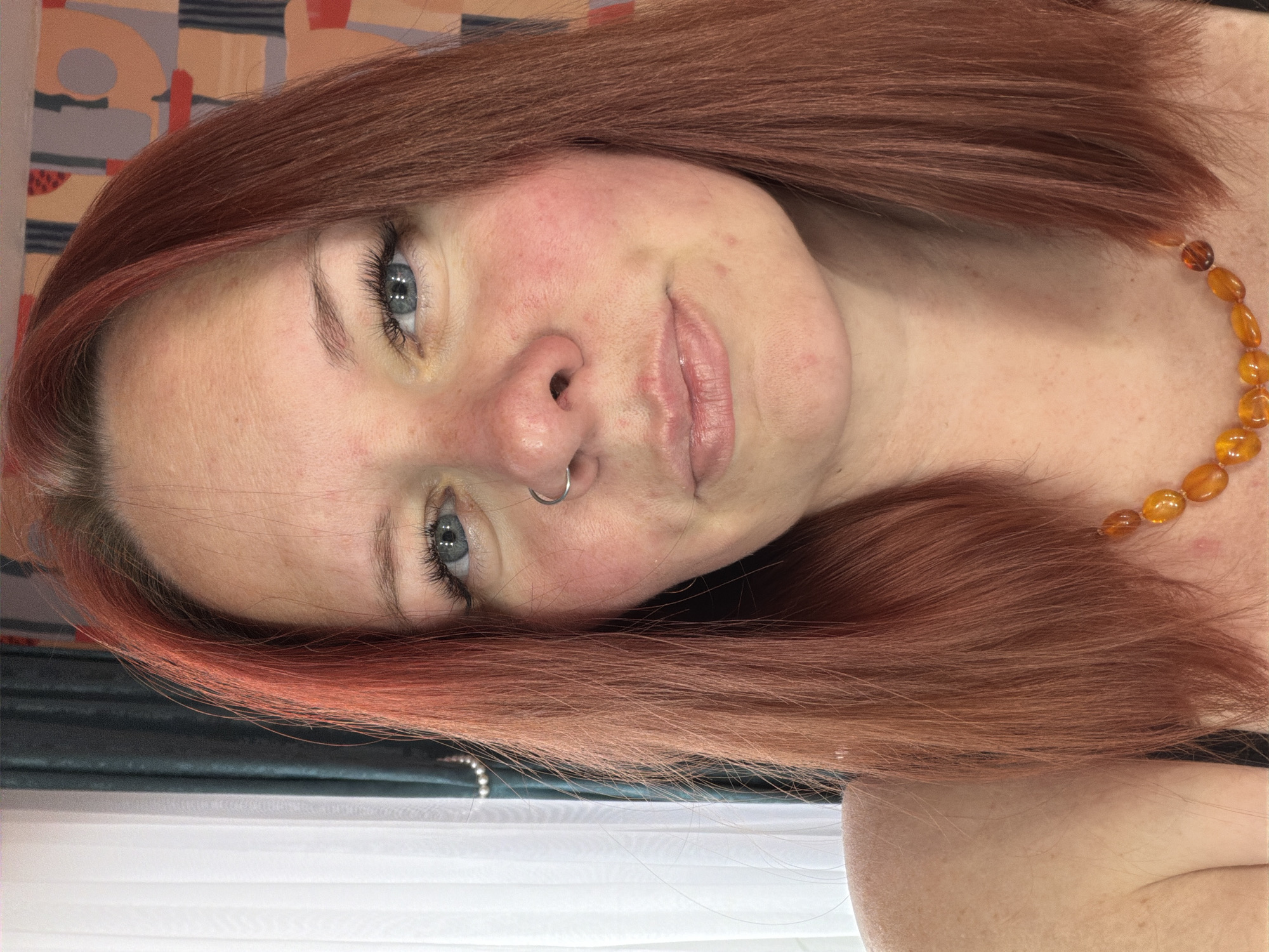 The image is a close-up, angled selfie of a woman. She has red-auburn hair styled with a slight wave, and it frames her face. Her eyes are light blue with dark lashes, and she has fair skin with some redness on her cheeks and forehead. She is wearing a nose ring on her left nostril. A necklace made of amber-colored beads is visible on her neck. The background is a mix of colors and shapes, suggesting an interior setting. The bottom of the image shows a sheer white curtain and a dark fabric. There is no information about the location being in Chaikovsky, Russia.
