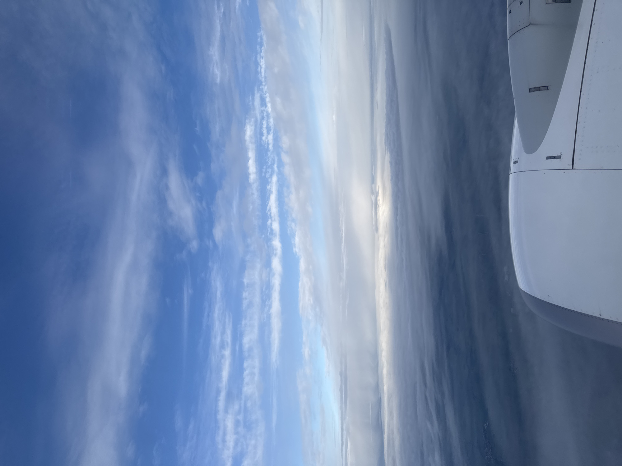 The image shows a view from an airplane window. The sky is visible with a mix of clear blue and white clouds, some in wispy formations. A portion of the airplane wing is visible in the right side of the frame. No specific location or people are visible.