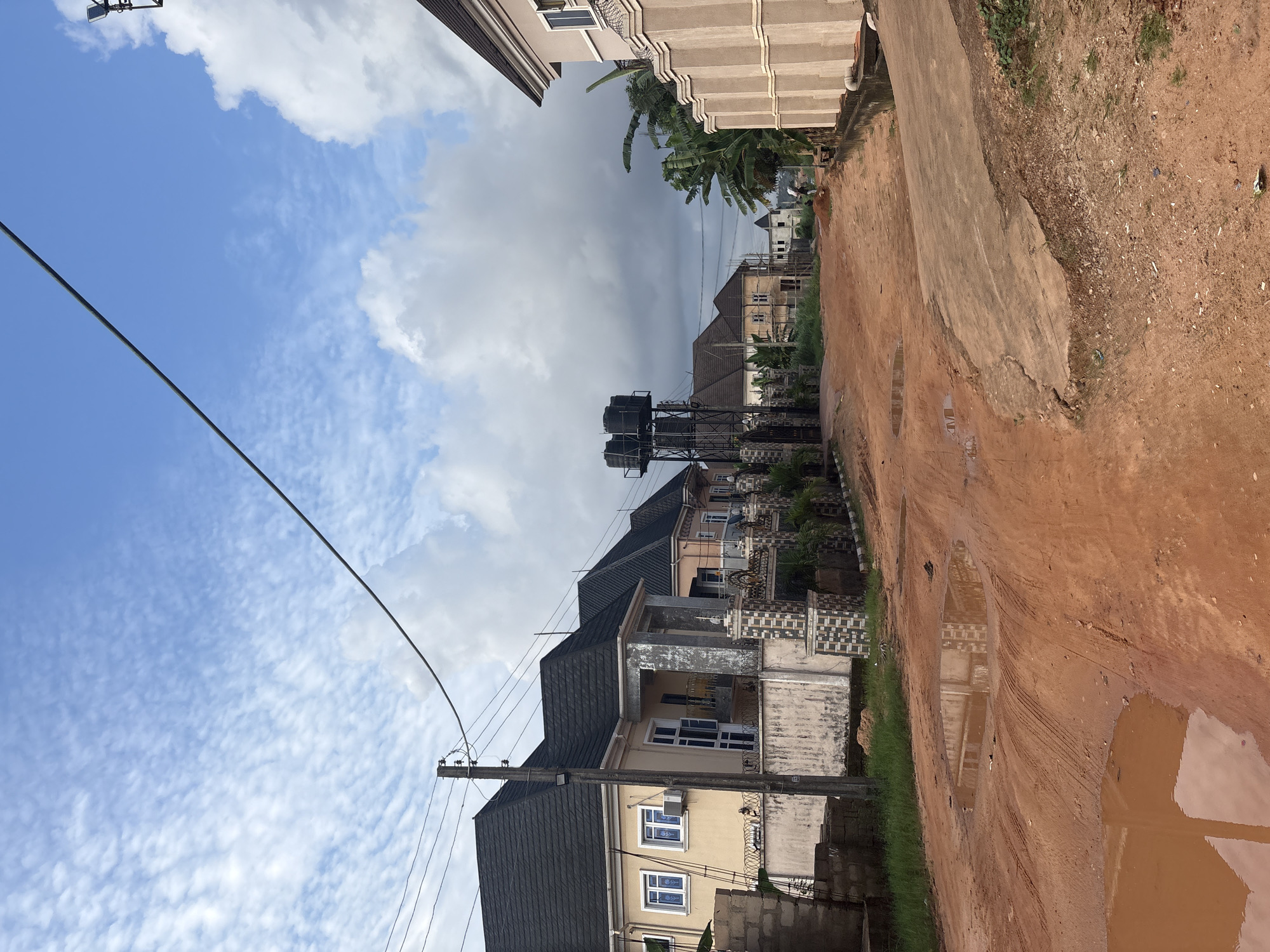 The image depicts a street in Obosi, Nigeria, characterized by its unpaved, reddish-brown dirt road with several large puddles reflecting the sky and surrounding structures. The road is uneven, with tire tracks visible in the mud.
On the left side of the road, several modern houses with dark tiled roofs are visible. These houses are painted in neutral colors and feature white-framed windows and air conditioning units. A concrete wall with decorative tiling and a metal gate separates one of the houses from the street. A utility pole with wires stretches across the top of the frame, originating from the left side.
Further down the street, a water tower with multiple tanks stands prominently. To the right of the water tower, more houses are visible, some appearing to be under construction with scaffolding.
The right side of the road is lined with a beige-colored building featuring a textured facade and a decorative wall. Banana trees and other lush greenery are present, suggesting a tropical or subtropical climate.
The sky is partly cloudy, with patches of blue sky visible through a layer of white and gray clouds. The overall impression is that of a residential area in Obosi, Nigeria, experiencing recent rainfall, as indicated by the puddles on the road. The scene appears calm and undeveloped, with no immediate signs of activity beyond the presence of the structures and vegetation.