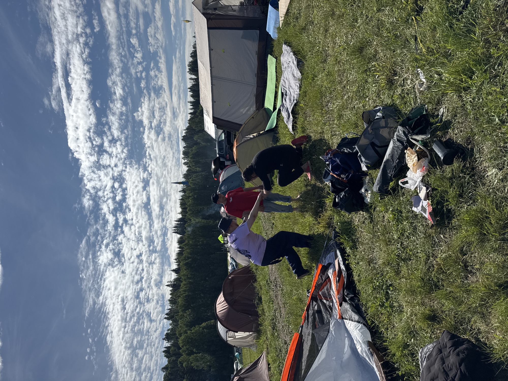 This image depicts an outdoor camping scene in Большая Кивара, Russia, under a partly cloudy blue sky. Three adult males are present in the midground on a grassy field. The man on the left, wearing a light purple t-shirt, dark pants, and a dark bucket hat, is in a dynamic posture, leaning forward with his left arm extended towards a crouching man. The crouching man, wearing a dark long-sleeved shirt and dark pants with red shoes, has his left hand extended towards the first man, suggesting a hand-to-hand interaction. A third man, wearing a red t-shirt and blue jeans, stands barefoot nearby, observing the interaction.
The immediate foreground shows camping equipment, including a partially disassembled tent (white, orange, black), multiple bags (dark blue backpack, grey duffel bags, black plastic bags), a black thermos or cup, and scattered paper or trash on the grass. In the midground and background, numerous tents of various sizes and colors are pitched across the field, including several dome tents (brown, beige, green) and a large, multi-room rectangular white tent with windows, in front of which green and blue sleeping mats are laid out. Two vehicles, a dark SUV and a blue car, are visible behind some of the tents. A small yellow flag on a pole is visible to the right, near the large white tent. The scene is bordered by a dense forest of coniferous trees in the background. Above the treeline, a dark, pointed spire with a blue base, possibly a church steeple, is visible in the distance. The overall lighting is bright, indicating a sunny day.
