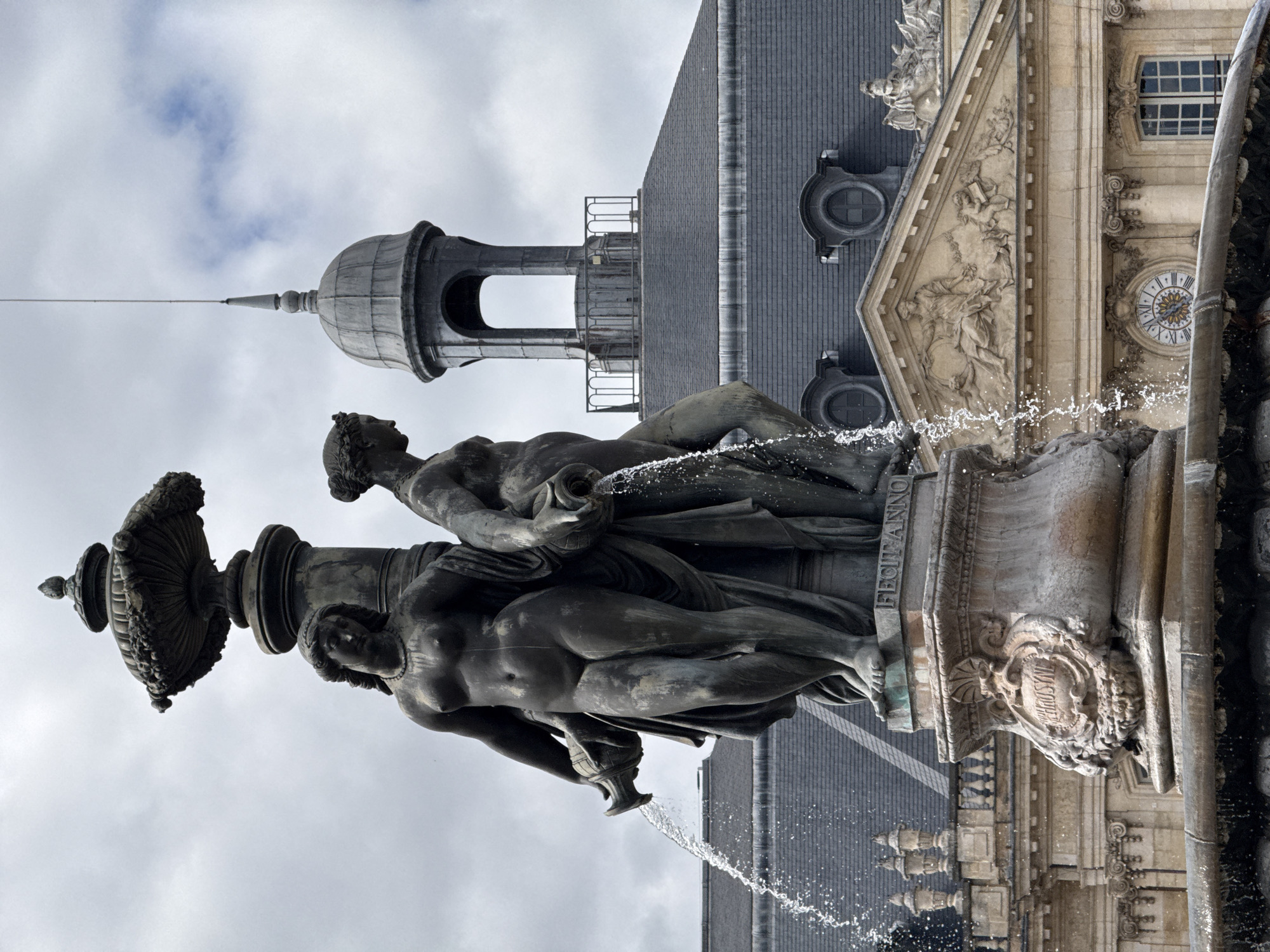 The image shows a detailed view of the Fontaine des Quatre-Sœurs (Fountain of the Four Sisters) located in the Place de la Bourse in Bordeaux, France. The fountain features three bronze statues of women pouring water from jugs, with water spraying outward. In the background, the elegant architecture of the Place de la Bourse is visible, including its distinctive grey mansard roof and ornate facade with a clock and bas-relief sculptures. The sky is overcast with clouds.