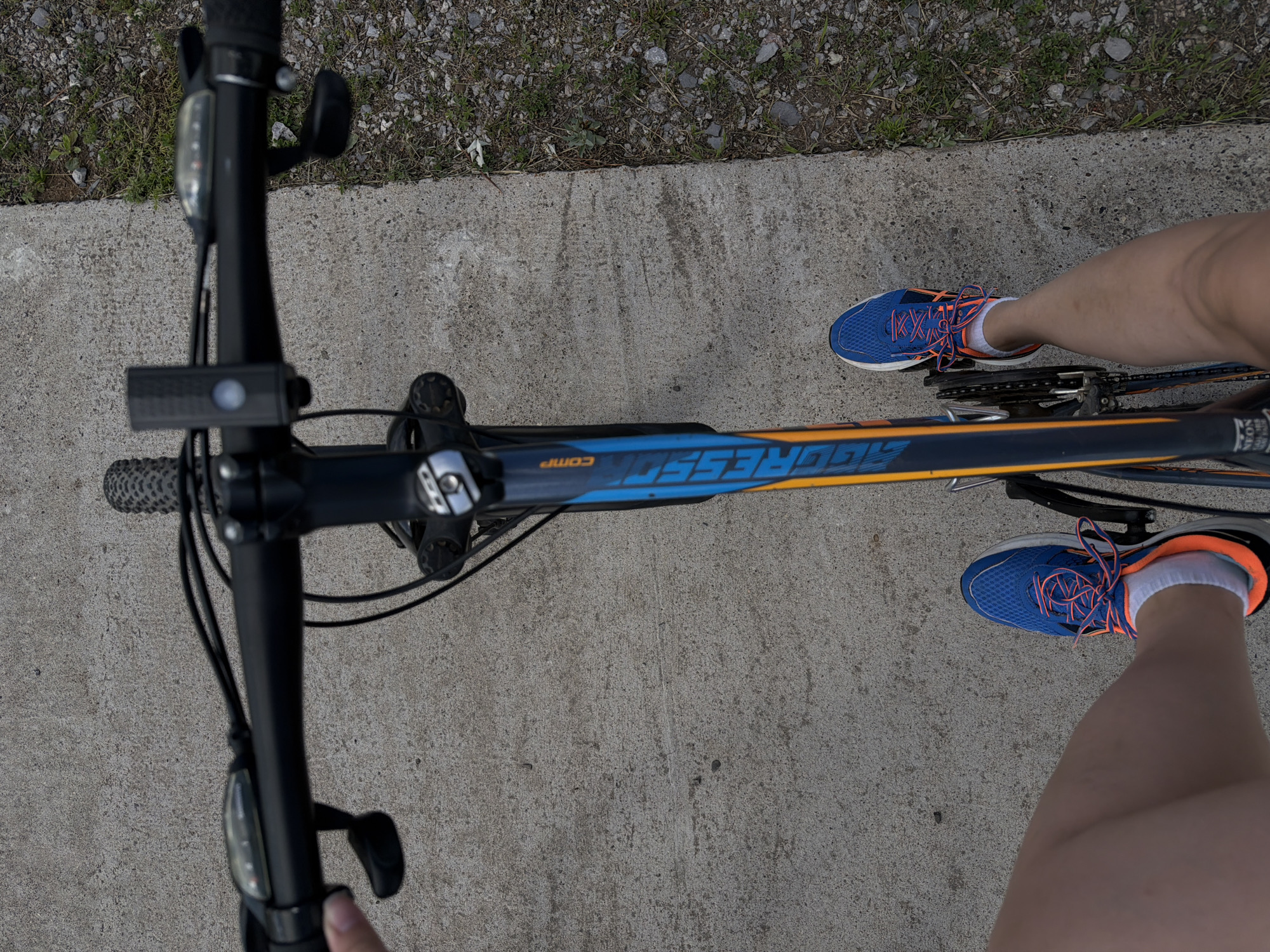The image shows a first-person perspective of someone riding a bicycle on a concrete path. The bicycle has a blue and orange frame with the brand "AGGRESSOR" visible. The rider is wearing blue athletic shoes with orange laces and white socks. A black handlebar with a light and gear shifters is in view. The path is bordered by gravel and sparse green vegetation on the right side. The scene appears to be an outdoor recreational area. The image does not provide any specific clues to pinpoint its location in Chaikovsky, Russia.