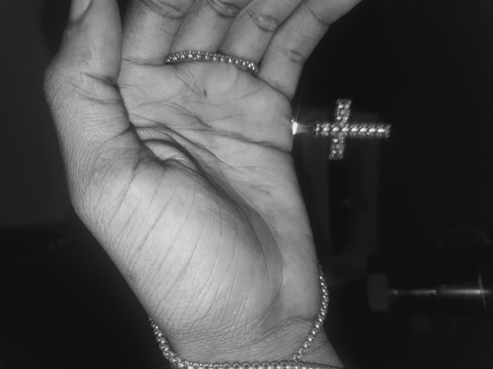 A black and white photograph shows a hand displaying jewelry. A ring with a beaded design encircles one finger, and a cross pendant is visible near the fingers. A beaded bracelet is on the wrist. The background is blurred and dark. The scene is not identifiable as Lokoja, Nigeria.