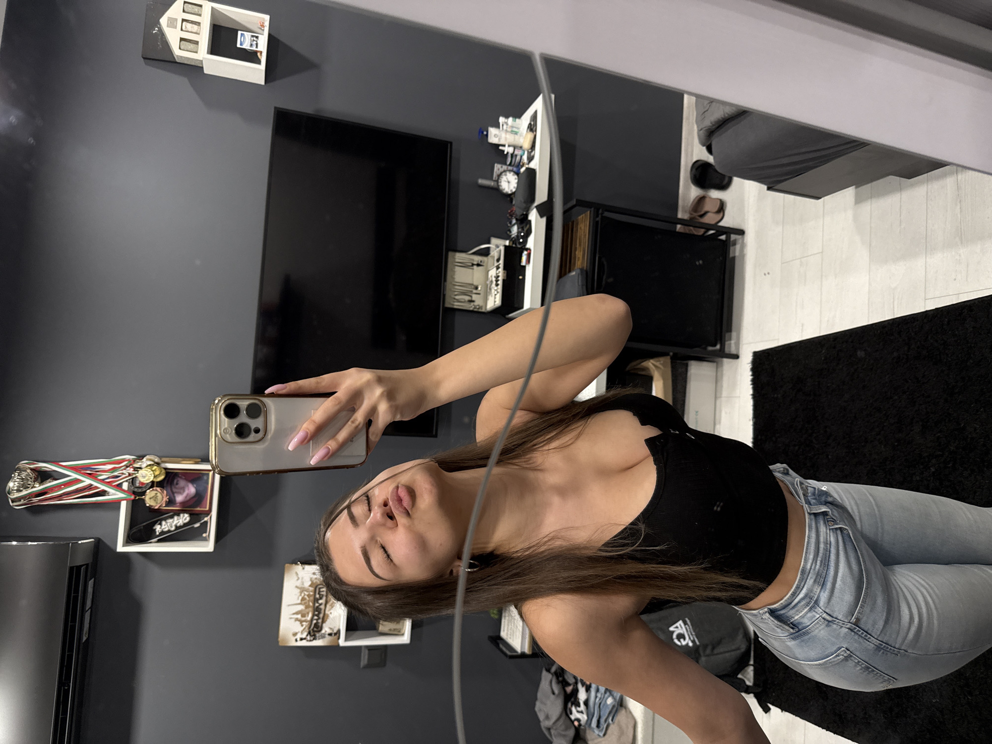 A young woman with long brown hair is captured in a mirror selfie, posing with puckered lips. She wears a black crop top and light blue jeans, holding a gold-cased smartphone with her right hand, revealing light pink manicured nails.
The setting appears to be an indoor room with dark gray walls and a light-colored floor, partially covered by a black shaggy rug. In the background, a large black flat-screen television is mounted on the wall. To the left, an air conditioning unit is visible.
Various personal items and decor adorn the walls and shelves. Above the television, a white house-shaped shelf contains small items, including a miniature picture that appears to show the letters "D_F". On the left wall, a white display shelf holds multiple medals with red, green, and white ribbons, alongside a framed picture and a miniature skateboard deck with "BADGIRL" written on it. To the right, another white shelf or table reflects an array of small items, including a watch and various bottles.
Further in the reflected background, a bed with gray bedding and some shoes are visible on the floor, suggesting a bedroom. A black bag with a white "KC" logo rests on the floor near the woman's leg. The scene captures a casual, modern interior with personal touches, likely taken during the day due to the ambient light, though no direct outdoor cues are present.
