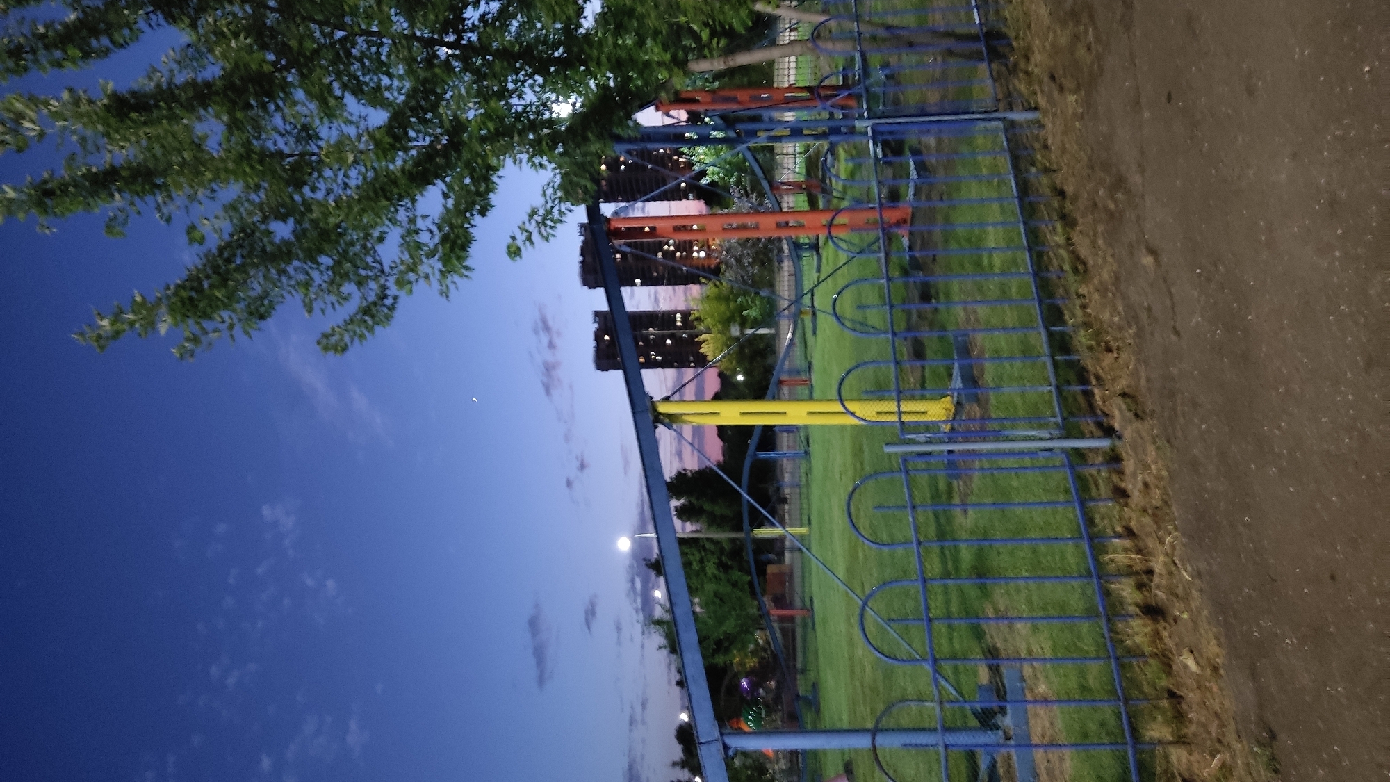 The image depicts an outdoor scene at dusk, likely in Krasnodar, Russia. In the foreground, a blue metal fence encloses a grassy area. Beyond the fence, colorful playground equipment, including orange and yellow poles, is visible. In the middle ground, several tall, modern buildings are silhouetted against a twilight sky. Trees line the edge of the grassy area, and a dirt path runs alongside the fence in the foreground. The sky is a deep blue, transitioning to hues of pink and orange near the horizon, indicating the sunset. A few wispy clouds are scattered across the sky, and a small, crescent moon is faintly visible. The overall mood of the image is peaceful and serene, capturing the transition from day to night. There is no visible text in the image.