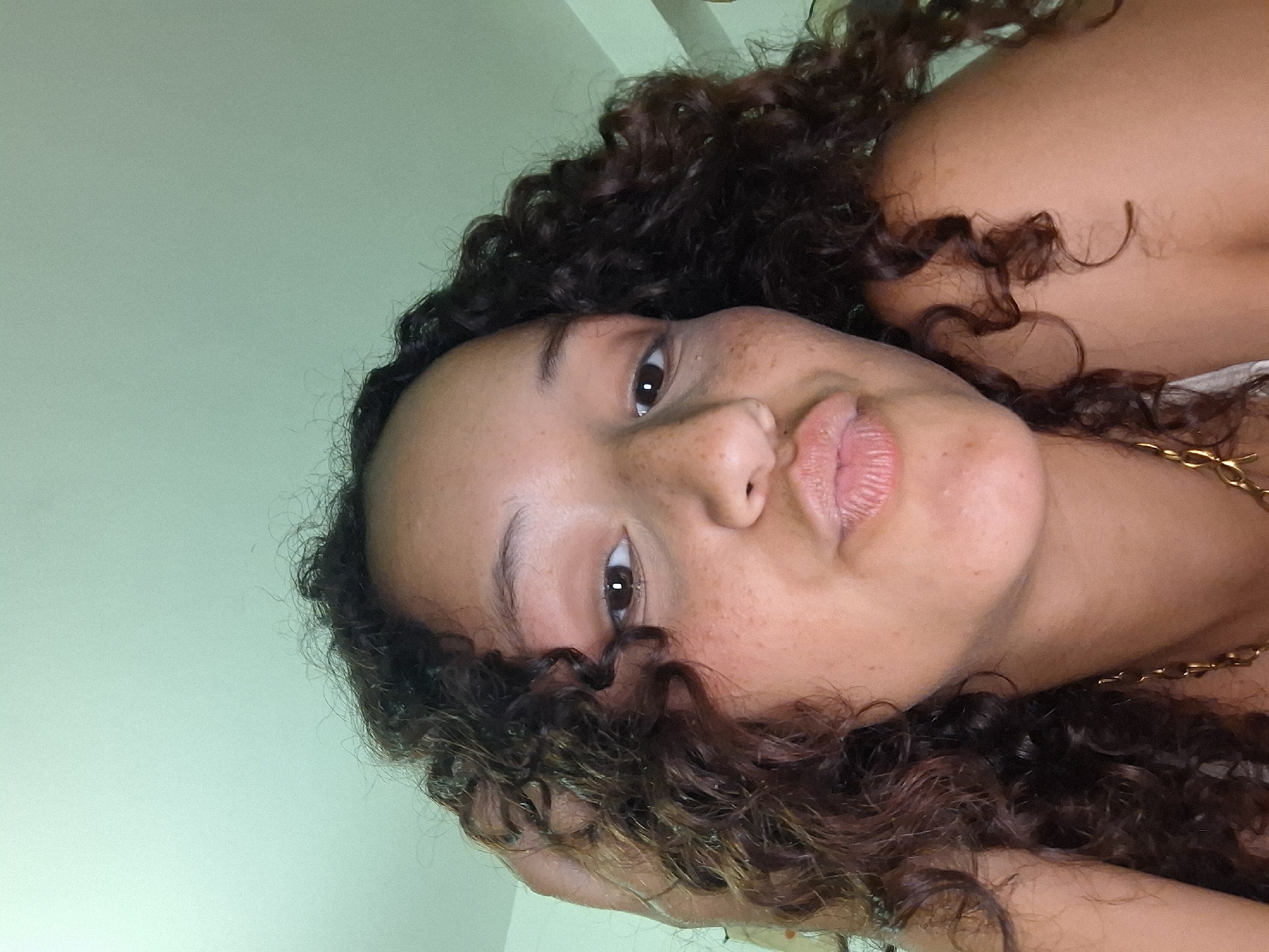 The image shows a close-up, low-angle, selfie-style photograph of a young woman with curly dark hair and fair skin with freckles. She is wearing a gold chain necklace. Her head is tilted slightly, and her right hand is resting on her hair, near her head. Her lips are pursed, giving the impression of a kiss. The background is a solid, light green color, suggesting an indoor environment. There are no discernible elements in the image that would allow for the identification of a specific location within Juan Griego, Venezuela.