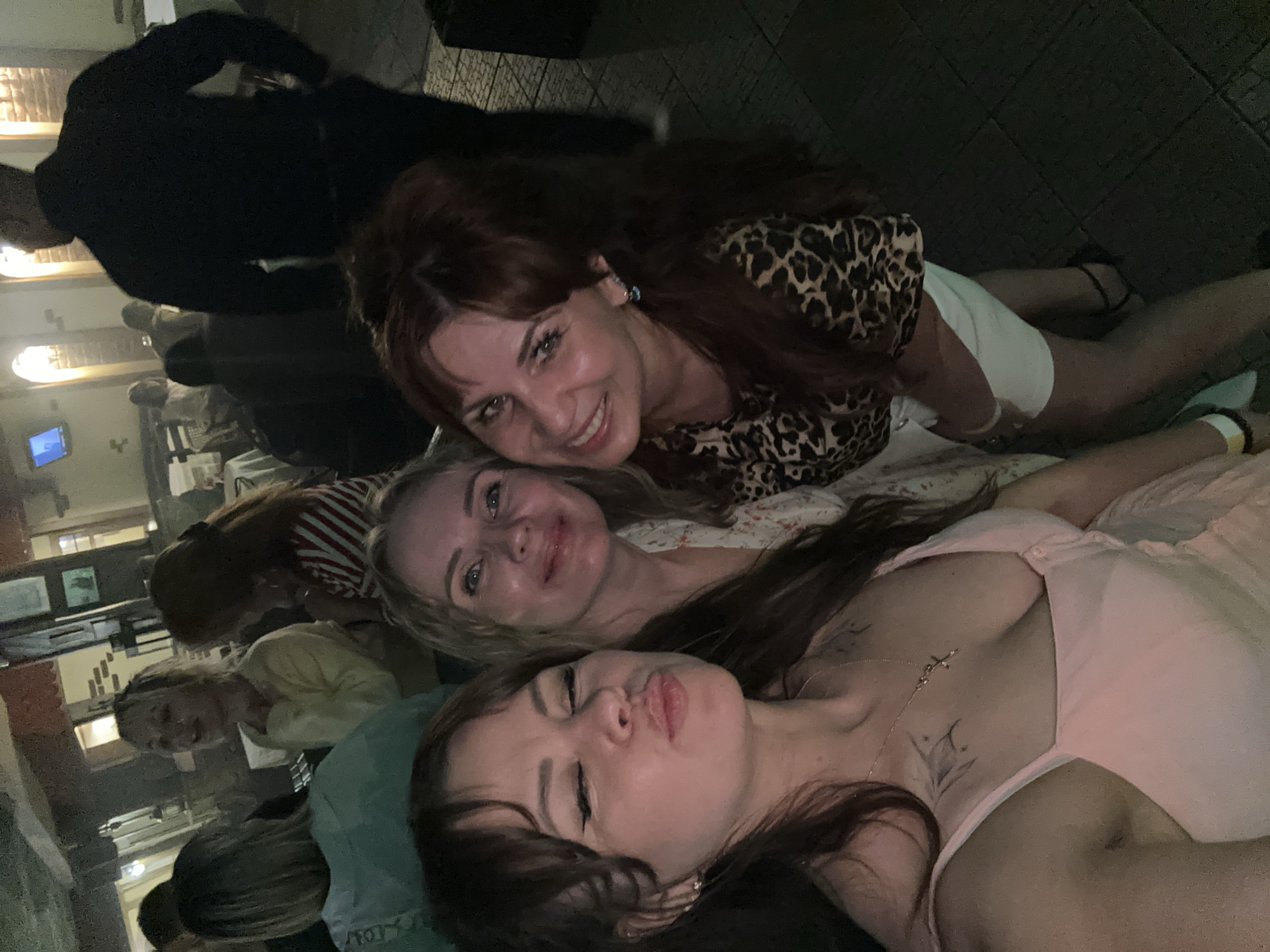 Three women are pictured in a selfie taken outdoors at night. The woman in the foreground on the left is wearing a pink dress and has her lips pursed. The woman in the middle is wearing a white floral dress and smiling. The woman on the right is wearing a leopard-print top and white shorts, and she is smiling. In the background, other people and a building can be seen. The scene appears to be a street or outdoor seating area of a restaurant or bar in Chaikovsky, Russia.