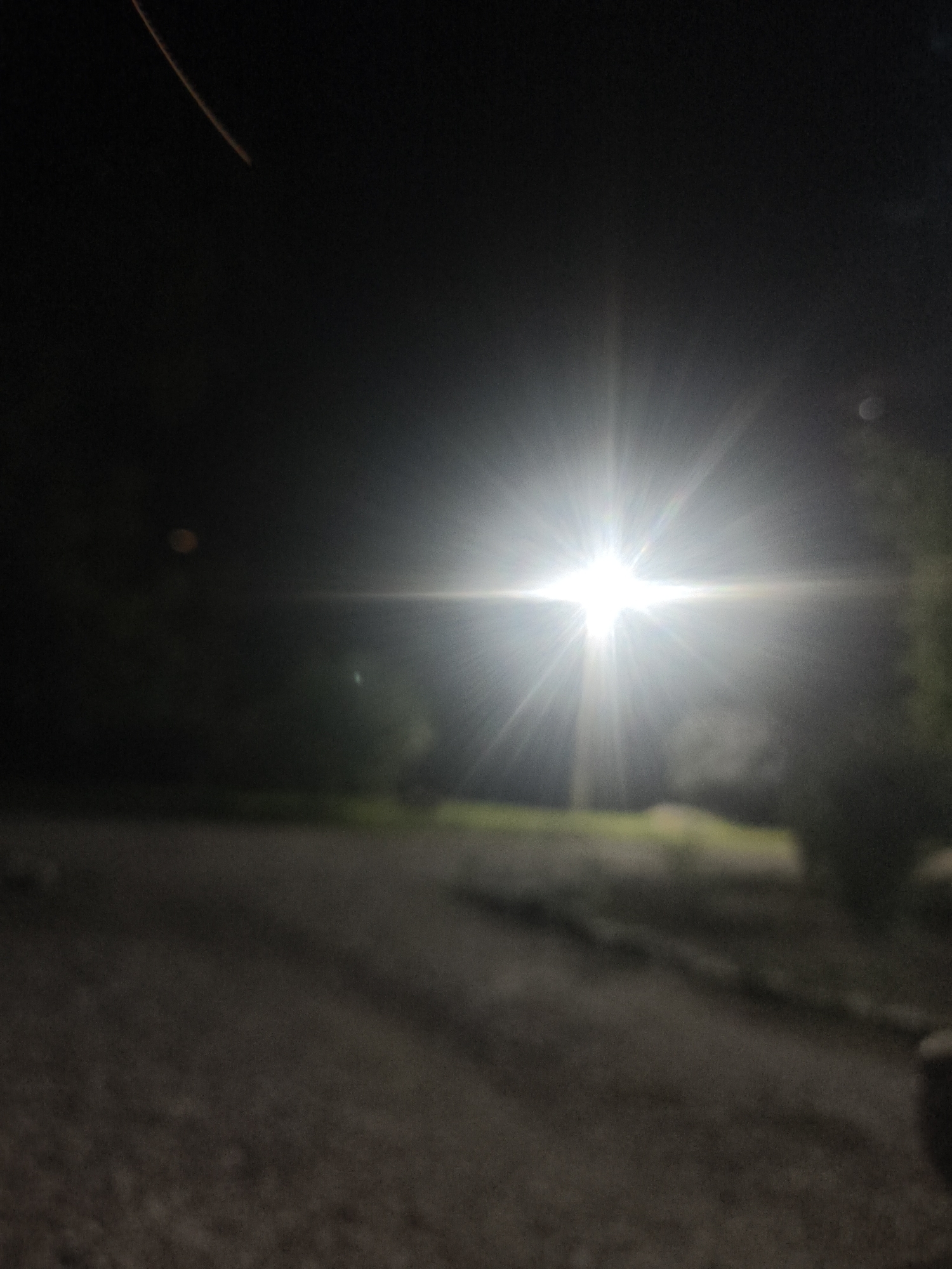 The image captures a nighttime scene with a strong light source, likely a street lamp or vehicle headlights, dominating the center. The light creates a starburst effect with prominent rays extending outwards. The foreground shows a dark, rough surface, possibly a road or pathway, with some indistinct shadows cast upon it. The background is dark and out of focus, with hints of vegetation or trees on the right and some blurred shapes that could be buildings or distant foliage. There is a faint, reddish streak in the upper left quadrant, possibly a lens flare or a light trail. No people or specific landmarks of Солдатская, Russia, are discernible in this image. The overall impression is one of low visibility and a stark contrast between the intense light and the surrounding darkness.