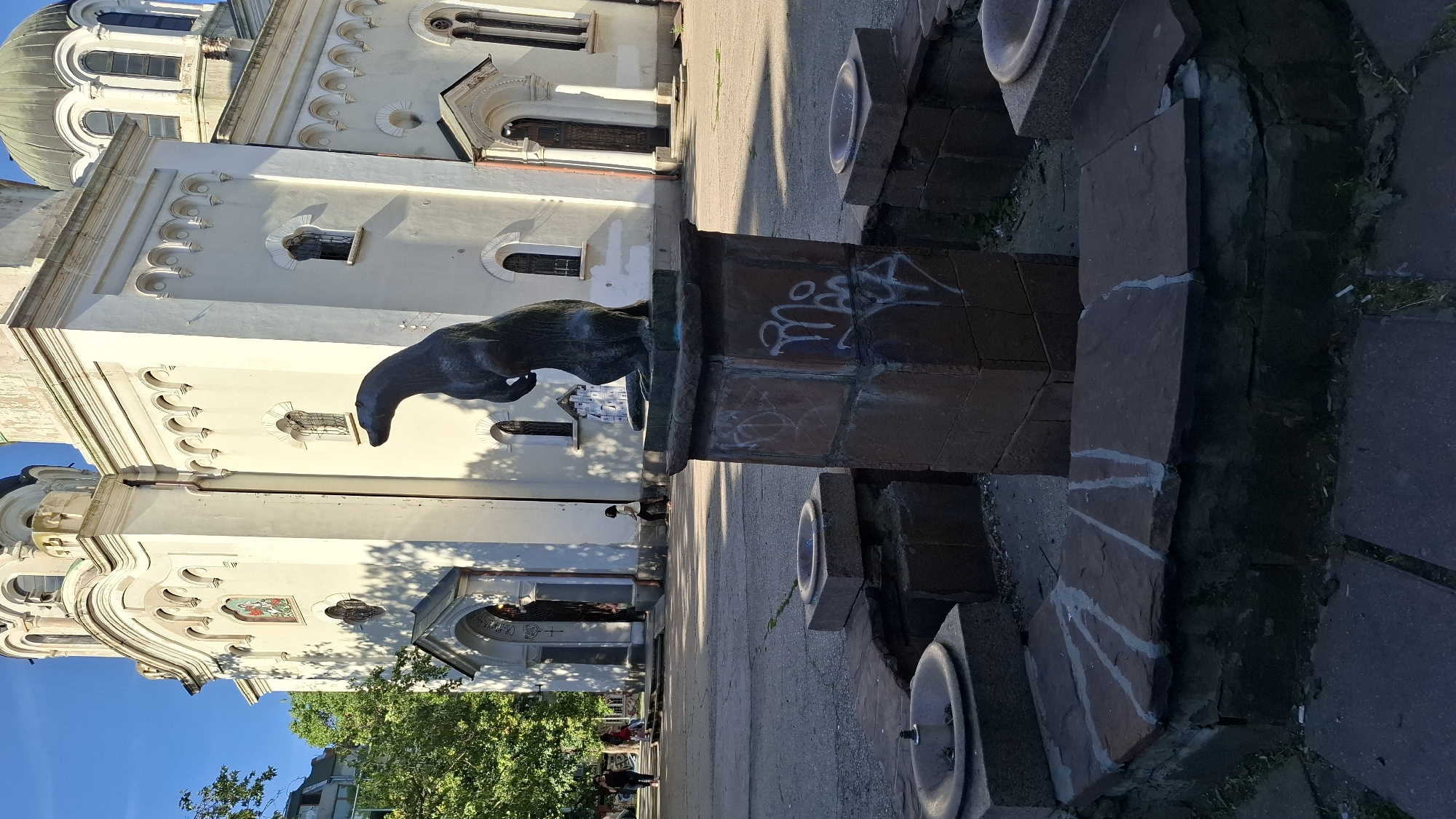 The image depicts a statue of a seal in front of a church in the city of Sofia, Bulgaria. The statue is made of dark material and stands on a stone pedestal that appears to be part of a dry fountain. The fountain has a rough, stone-lined basin. Graffiti is visible on the pedestal. The church, which has white walls and a green dome, is the backdrop. The sun casts shadows on the church and the surrounding paved area. Several people are visible in the background, walking and standing in front of the church entrance. Trees with green foliage are also present. The sky is blue and clear.