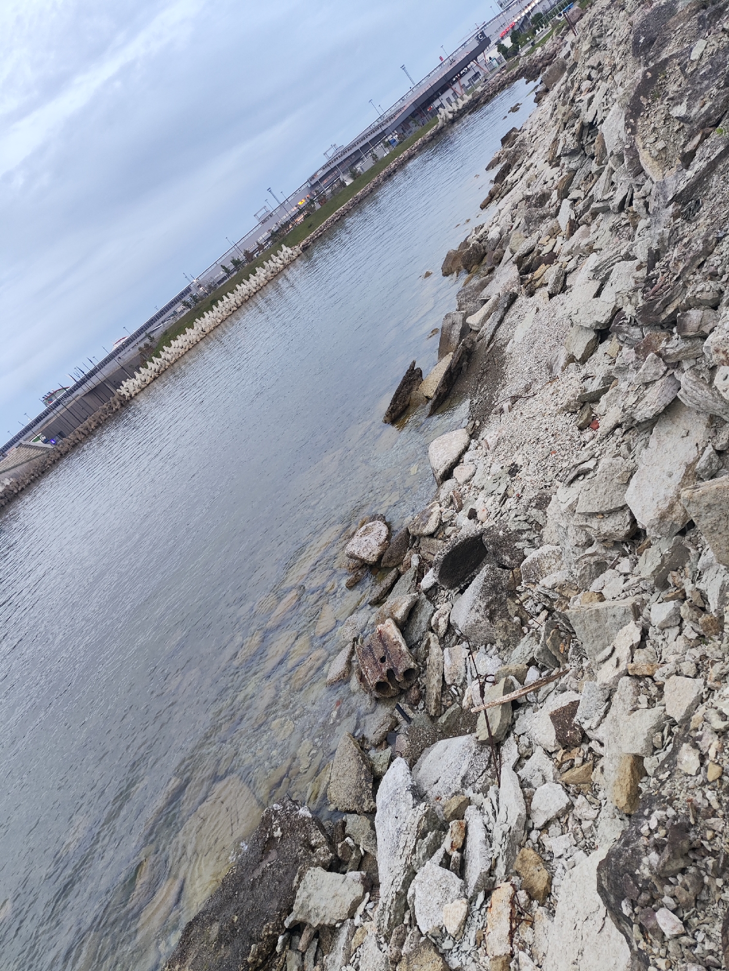 The image depicts a waterfront area with a rocky, debris-strewn shore and calm, grey water. In the background, a long, elevated structure, likely a bridge or elevated highway, runs parallel to the coast, with buildings and infrastructure visible beyond it. The sky is overcast with clouds. The scene appears to be an industrial or port area, possibly undergoing construction or renovation, given the presence of scattered concrete and rubble along the water's edge. The overall impression is one of an urban coastline with a utilitarian and somewhat neglected appearance.