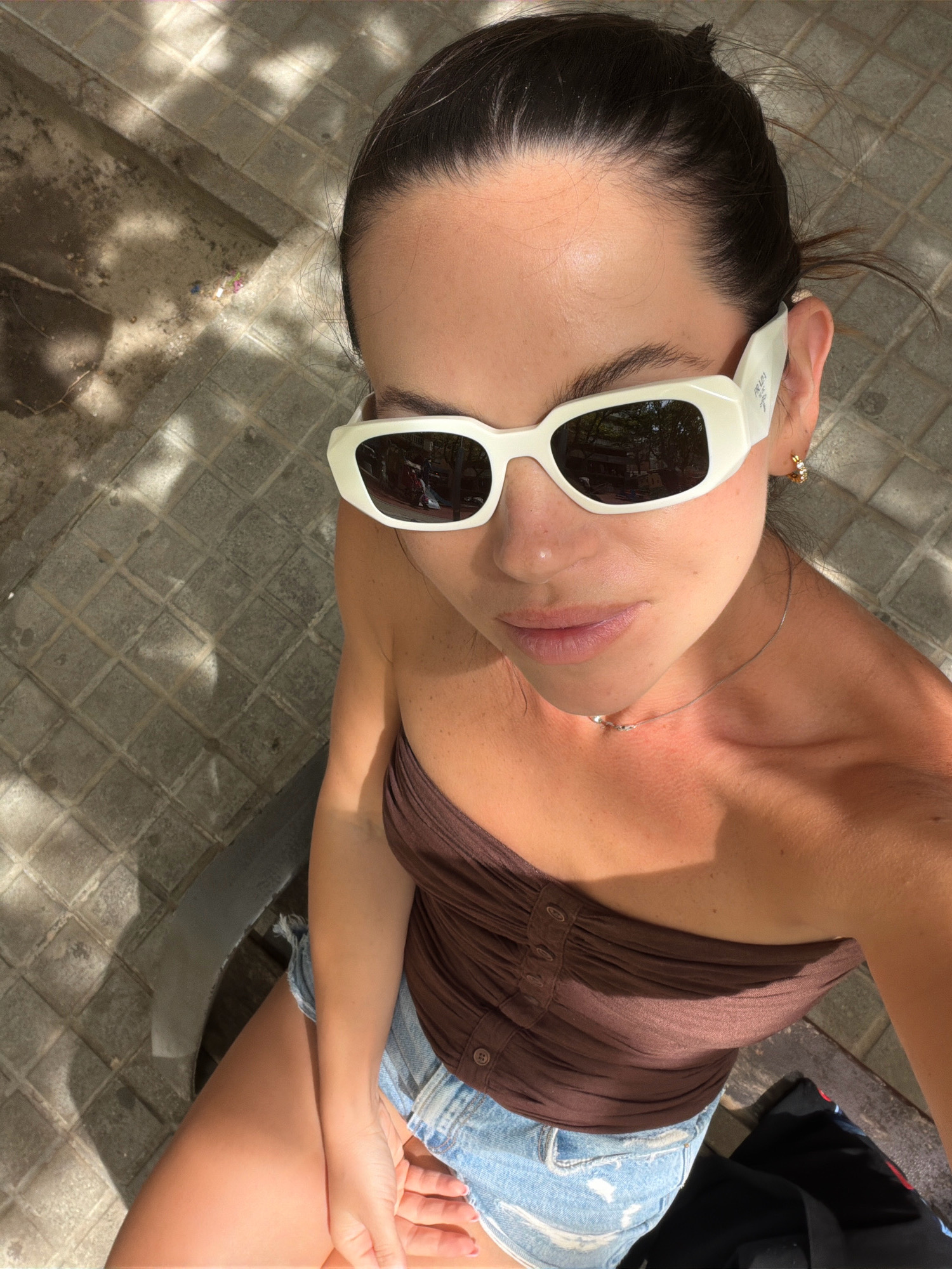 A young woman is shown from a high angle, seated outdoors on a sunny day. She has dark hair pulled back and is wearing large white-framed sunglasses. Her outfit consists of a brown strapless top and denim shorts. She is wearing a delicate silver necklace and a small hoop earring on her right ear. The background shows a paved area with square tiles, dappled with sunlight and shadow. A hint of a bench is visible beneath her. The scene appears to be a casual outdoor setting, possibly a public space like a park or plaza, within the city of Salou, Spain, given the architectural style of the paving stones which are common in coastal Spanish towns.