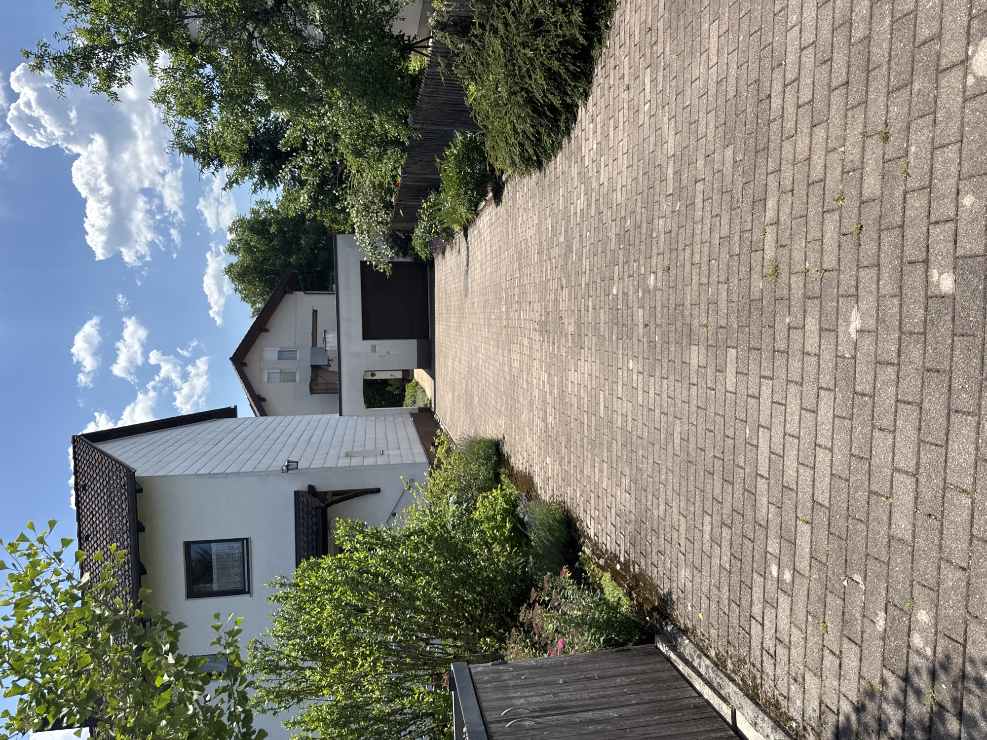 The image shows a paved driveway in front of a residential property. The driveway is made of interlocking brick pavers and is bordered by green vegetation and a wooden fence. A white house with a dark tiled roof is visible on the left side of the image, and another building with a dark garage door is visible at the end of the driveway. The sky is blue with white clouds. The location is identified as Höhenkirchen-Siegertsbrunn, Germany. There are no people or animals visible in the image. The scene appears to be a typical residential street on a sunny day.