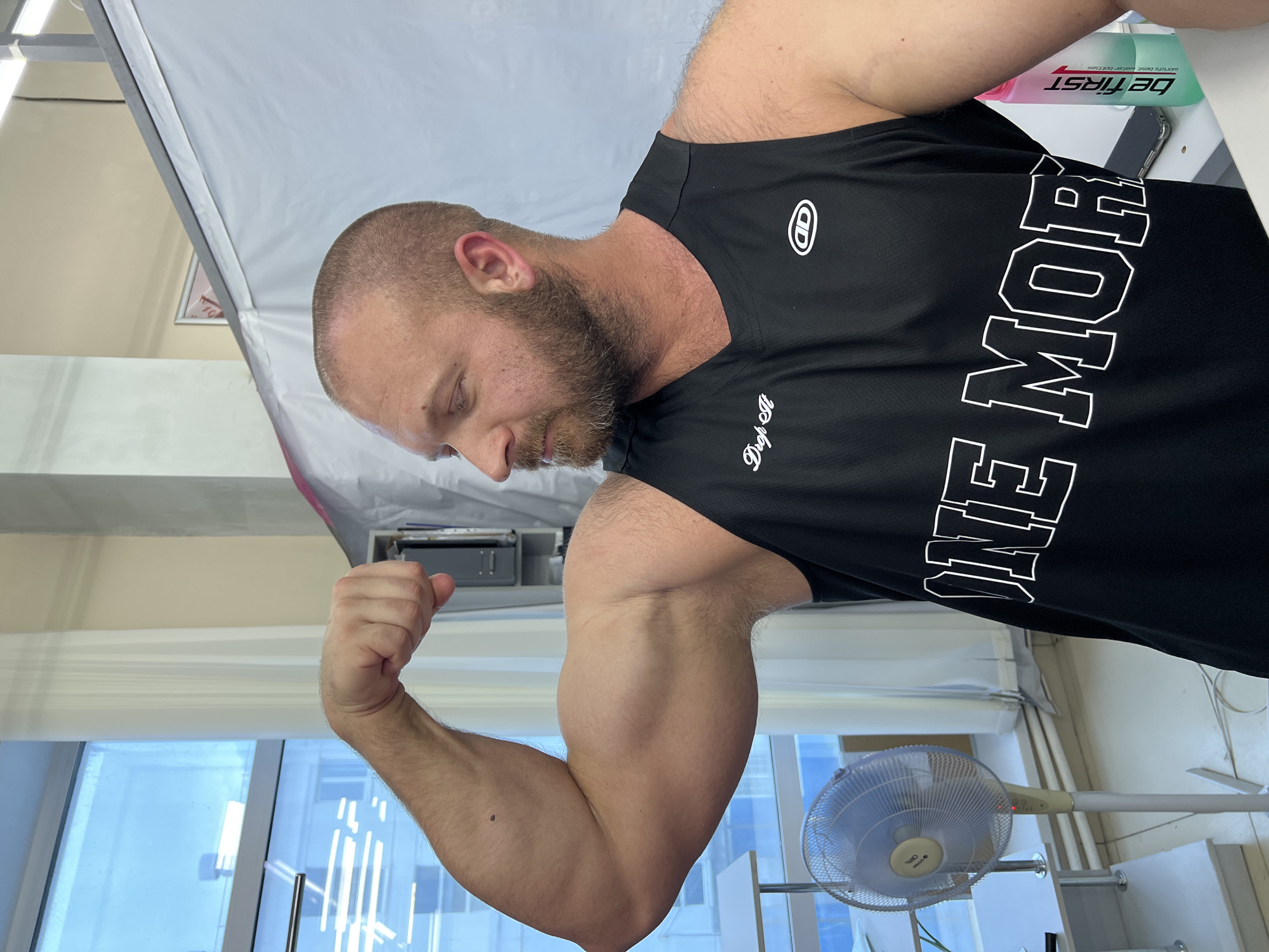 A man with a shaved head and beard flexes his bicep in an indoor setting, likely a room or studio. He is wearing a black tank top with white lettering that reads "ONE MOR." A floor fan is visible to his left, and a window with white curtains is behind him. A pink and green water bottle with the label "be first" is also present. The overall environment appears utilitarian. There is no indication of the specific location within the city of Chaikovsky, Russia.