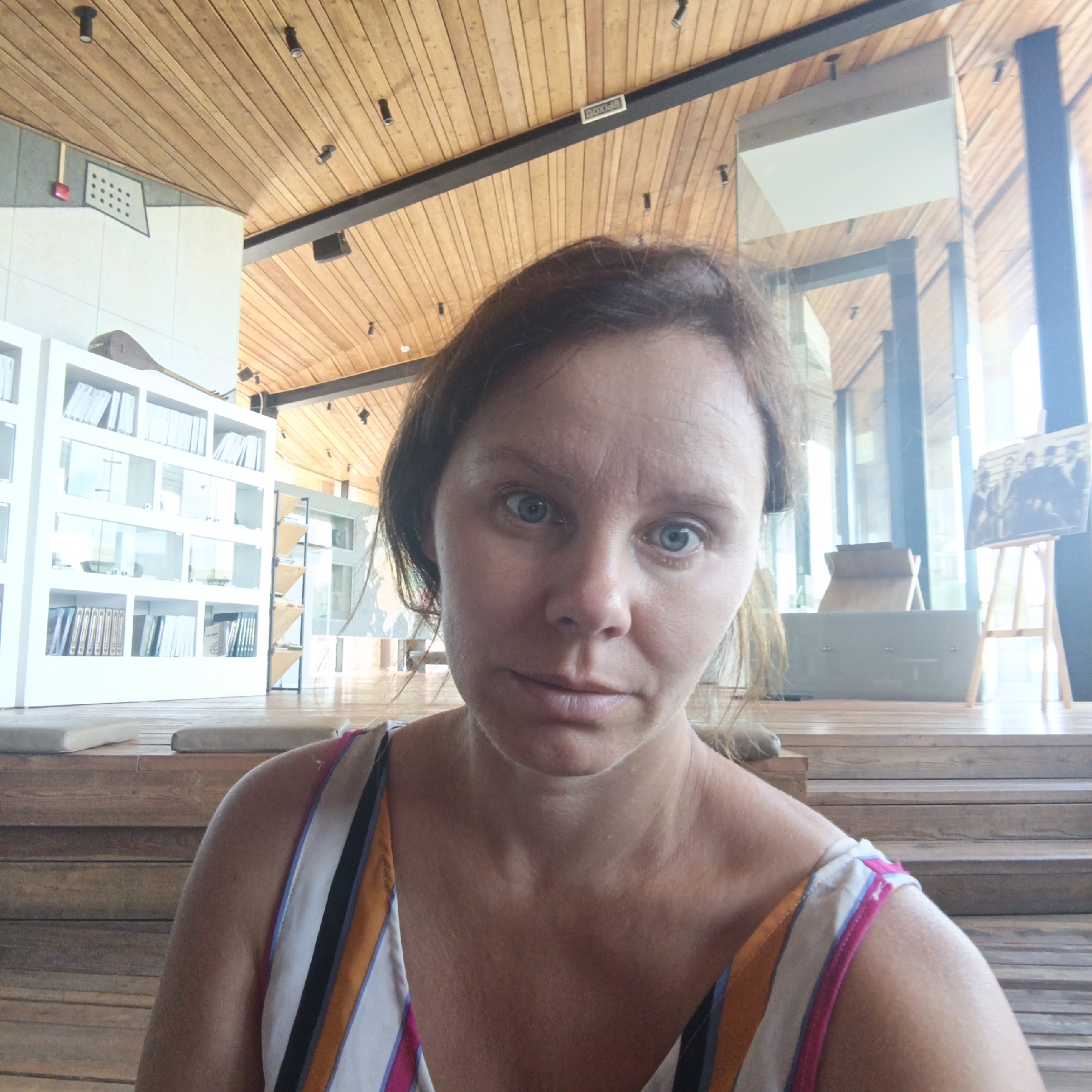 The image is a selfie of a woman with light skin and medium brown hair, taken from a low angle. She has blue eyes and appears to be indoors. She is wearing a colorful striped top with orange, white, pink, and black stripes. The background shows a modern interior with wooden paneling on the ceiling and wooden stairs. To the left, there is a white bookshelf filled with books and some decorative items. A painting of a group of people is displayed on an easel to the right. There is no indication in the image that this location is in Verkhnie Termy, Russia.