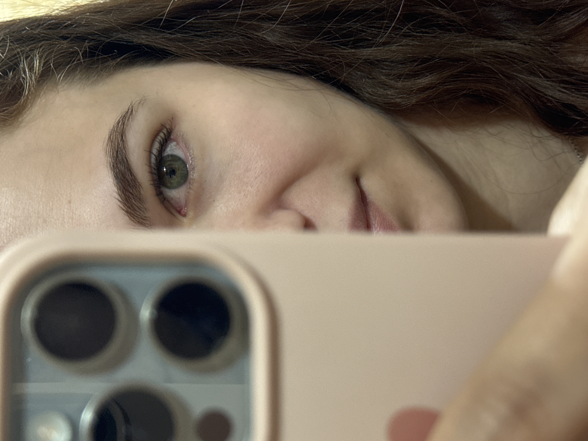 A close-up photograph shows a woman taking a selfie. Her face is partially visible, with one green eye and part of her smile visible. Her brown hair is visible above her forehead. The camera being used is a smartphone with a pink case, featuring three prominent camera lenses. The background is out of focus. There is no indication of the location being Nizhnyaya Gar', Russia, or any environmental context beyond what is immediately visible.