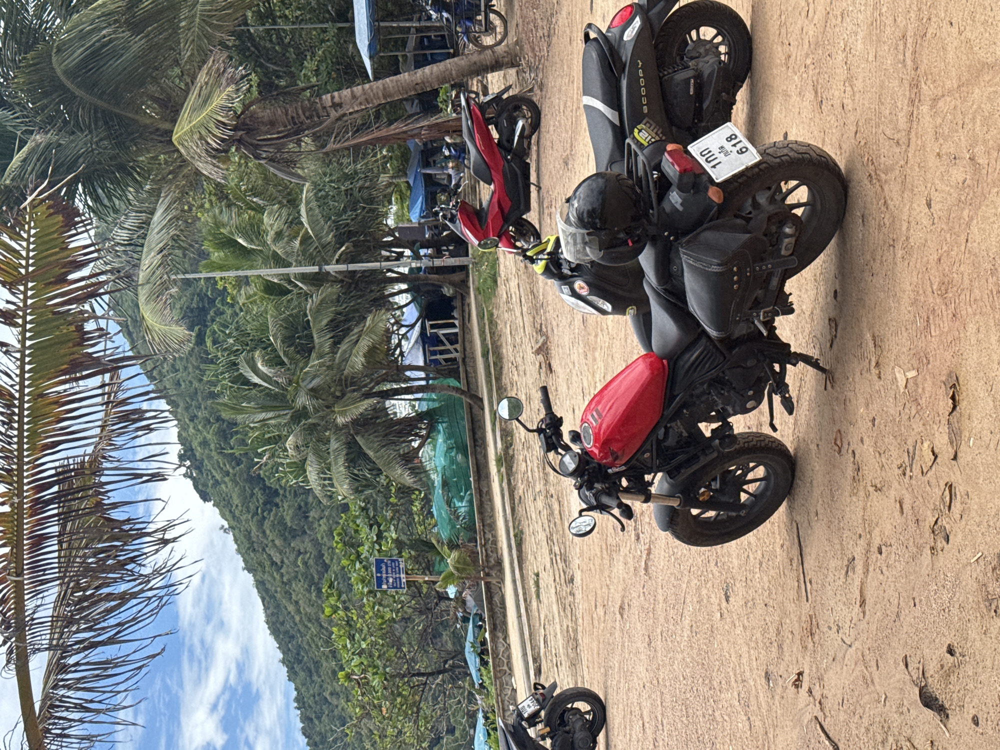 The image depicts a scene on a sandy area in Rawai, Thailand, with several motorcycles parked. In the foreground, a red motorcycle with leather saddlebags and a black helmet is prominently displayed, its license plate reading "100 กทม 618". Behind it, a black scooter with the brand name "SCOOPY" is visible. Further back, another red scooter is parked, and a black motorcycle is seen on the left. The background features lush green mountains, palm trees, and vegetation, with several blue tents and umbrellas suggesting a beachside location. The ground is covered in sand and some debris, indicating an outdoor environment. The sky is partially cloudy, with visible clouds and blue sky.