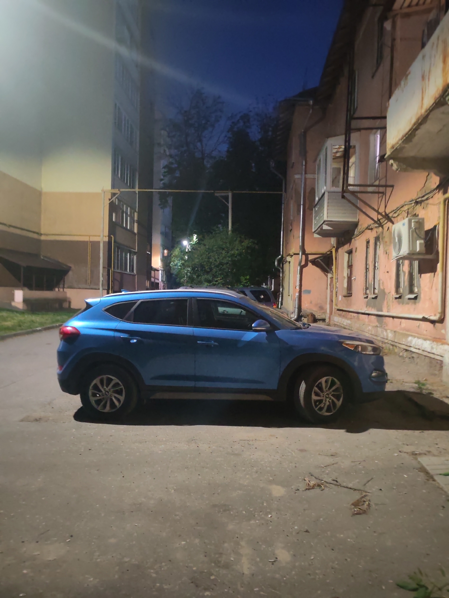 A blue Hyundai Tucson is parked on a paved street at night, illuminated by streetlights. The car is positioned in the middle of the frame, facing left, with its rear end slightly angled towards the viewer. The street is flanked by buildings: to the left, a tall, modern apartment building with multiple windows; to the right, an older, multi-story building with a textured, peach-colored facade, balconies, and visible external pipes and air conditioning units. A few trees are visible behind the buildings. The sky above is dark blue, indicating nighttime. There are no people or animals in the frame. The scene appears to be an urban residential area.