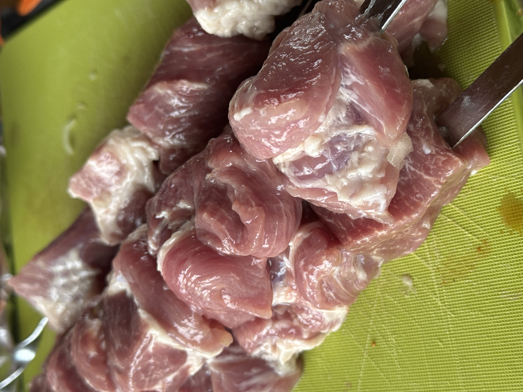 A close-up, high-angle shot captures numerous chunks of raw, reddish-pink meat, likely pork or lamb, skewered on metal rods. The meat pieces, varying slightly in size, exhibit streaks of white fat and connective tissue, appearing moist and fresh. Two metal skewers are visible, one prominently piercing several cubes of meat, while another is partially seen on the left side of the frame. The meat rests on a bright lime-green surface, which appears to be a textured cutting board or tray with a distinct grid pattern. Some faint, lighter marks or liquid residue are visible on the green surface, possibly from the meat preparation. The focus is sharp on the meat and skewers, with the background slightly blurred. The scene suggests the preparation of shashlik, a popular dish in Russia, consistent with the provided location context of Krasnoyarsk. No people or text are visible in the image.