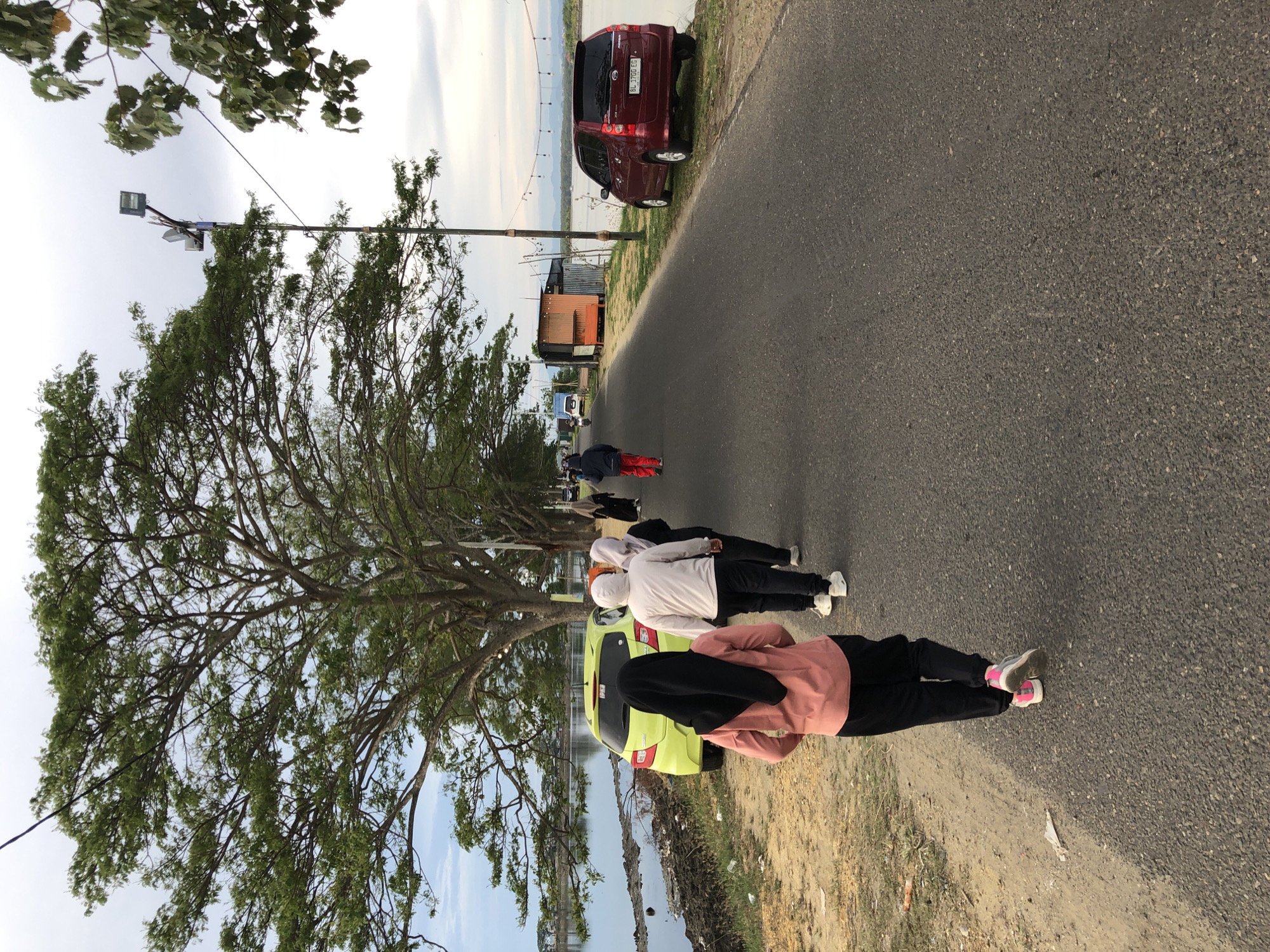 In Lhokseumawe, Indonesia, along a paved road bordering a body of water, four individuals are walking away from the camera. The person in the foreground is wearing a coral-colored long-sleeved shirt and black pants with pink and grey sneakers. Behind them, three other people are walking in the same direction. One is wearing a light-colored hooded jacket and black pants, another a white hooded jacket and black pants, and the third is wearing a dark headscarf and black attire. A bright green hatchback car is parked on the left side of the road, near the water. Further down the road, a dark red minivan is parked on the right. The environment features a line of large trees with full canopies lining the left side of the road and a cloudy sky overhead. A street lamp with multiple light fixtures is visible on the right side of the road. Small structures resembling food stalls or small buildings are present further down the road on the right.