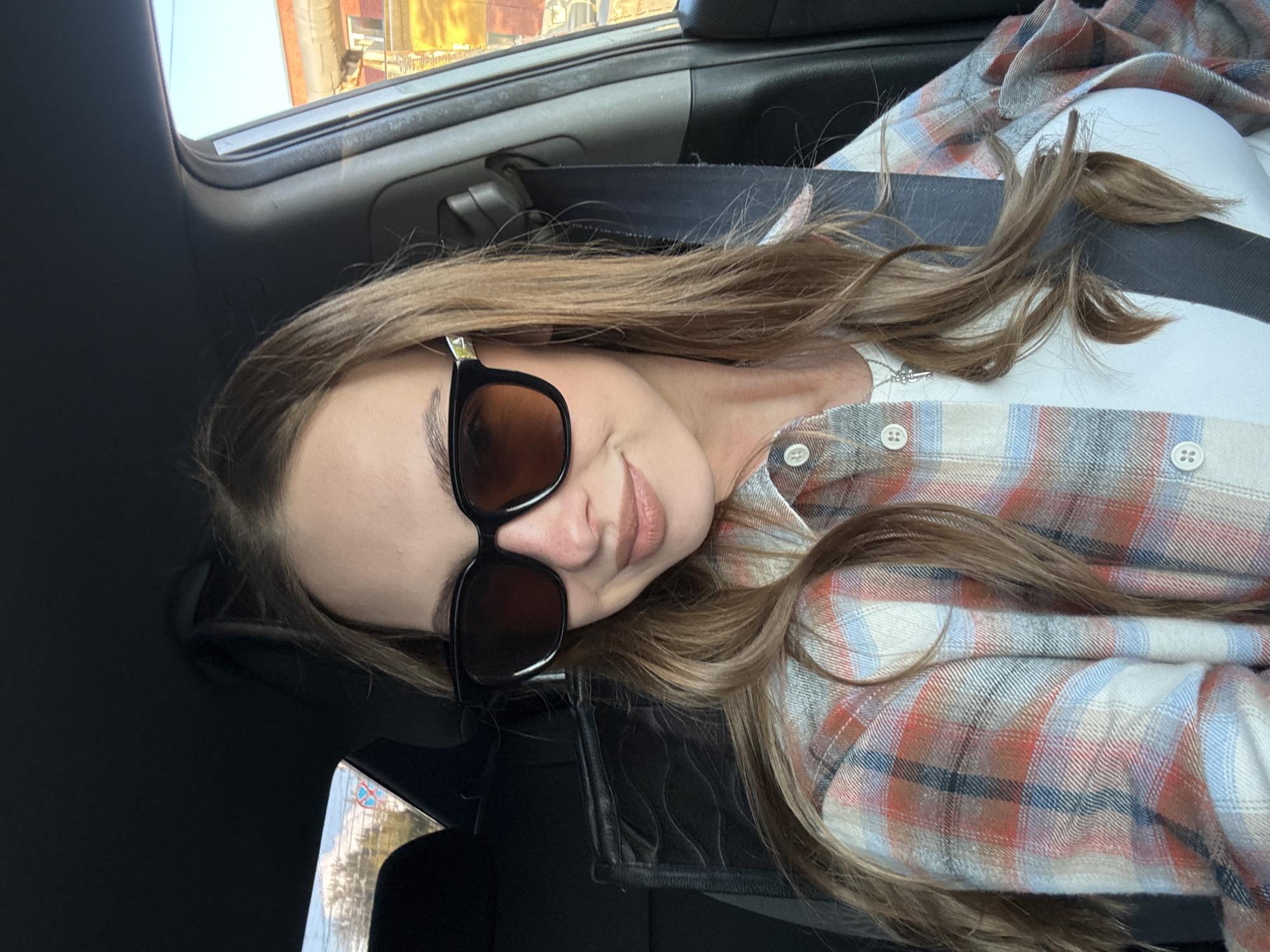 A young woman with long, brown hair and light skin is in the driver's seat of a car. She is wearing large, dark-rimmed sunglasses, a white shirt, and a red, white, and blue plaid flannel shirt. Her hair is partially visible and falls over her shoulders. The woman is looking at the camera and smiling slightly. The interior of the car is visible, including the dashboard, steering wheel, and seatbelt. The background outside the car window shows buildings and trees. The lighting suggests it is daytime.