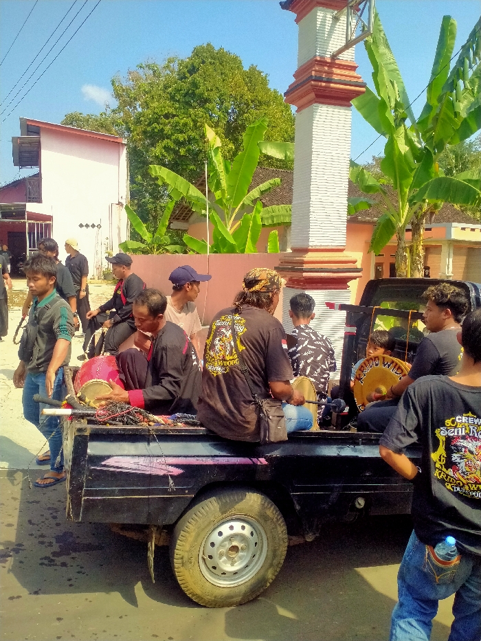 The image captures an outdoor daytime scene in Tawangsari, Indonesia, featuring approximately 10-12 adult males gathered around a black pickup truck. Several men are seated within the truck's bed, which holds traditional musical instruments. One man, wearing a black long-sleeved shirt, cradles a red drum (kendang). Another man, seen from behind, wears a black T-shirt with a yellow and white design and text "SENI RA[...] KRIDO WIDODO," and has a patterned bandana on his head; he holds a small, round, possibly metallic instrument with a beater. A third man in the truck also holds a similar instrument, displaying the text "KRIDO WIDODO." Other musical equipment, including ropes, is visible in the truck bed.

To the immediate left of the truck, a man in a dark polo shirt and blue jeans stands with his hands near his sides. Another man, partially visible from behind on the far right, wears a black T-shirt with a similar "CREW SENI RA[...] KRIDO WIDODO" design and blue jeans, with a plastic water bottle tucked into his rear pocket. Other men are visible in the background, to the left of the truck.

The environment includes a prominent tall, white, and red painted concrete pillar or monument on the right. In the background, residential buildings are visible, including a large, light pink/white structure with a red roof on the left, and a smaller, light orange building on the right. Lush green banana trees and a large deciduous tree are also present. The ground is paved, exhibiting dark stains. The sky is clear blue, indicating bright, sunny weather.