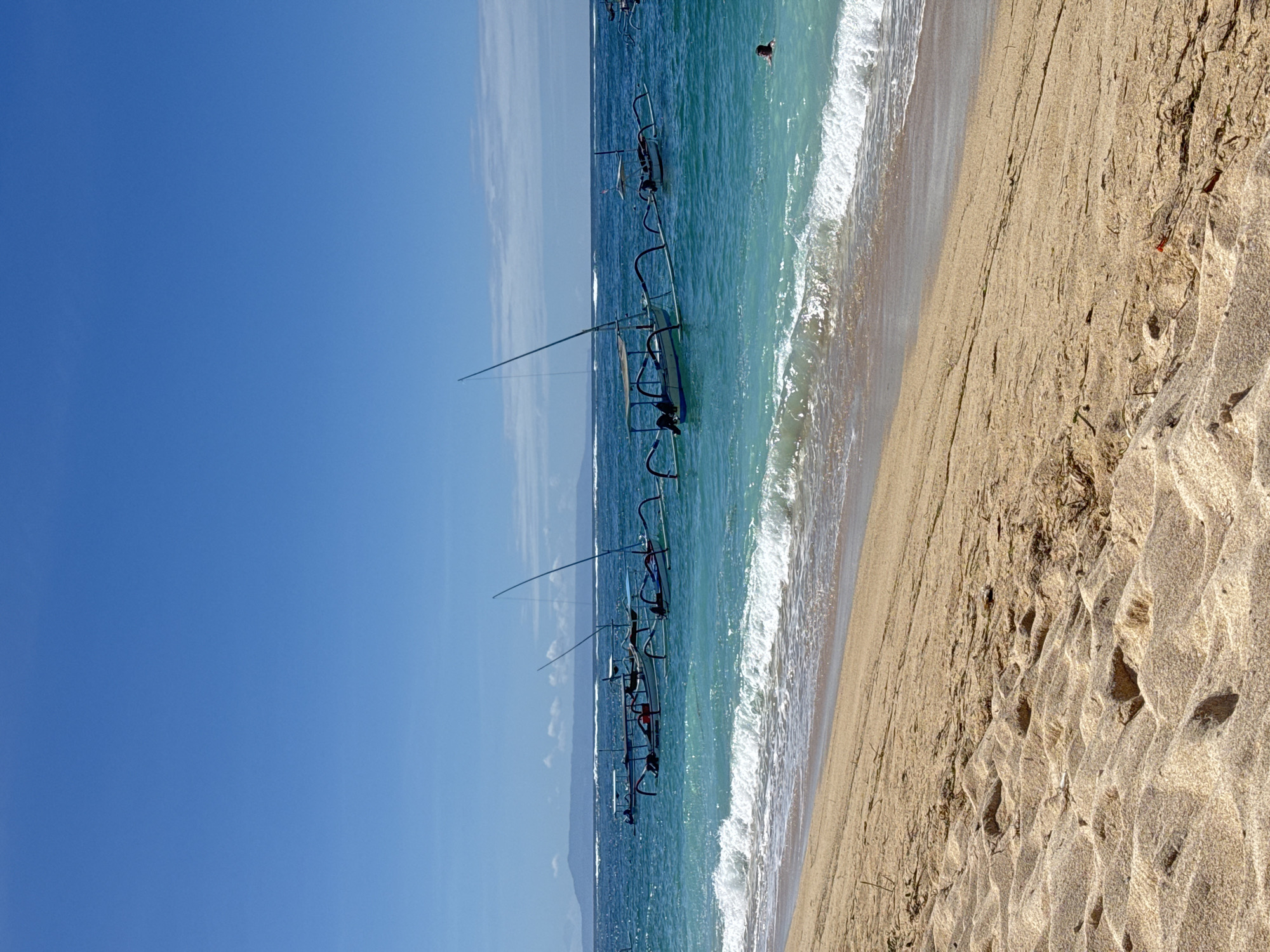 A beach scene shows several boats in the water. The boats are narrow and each have a long mast. The sand is light brown and there are wave patterns present in the water. The sky is a clear blue. The location is in the city of Unknown, Unknown.