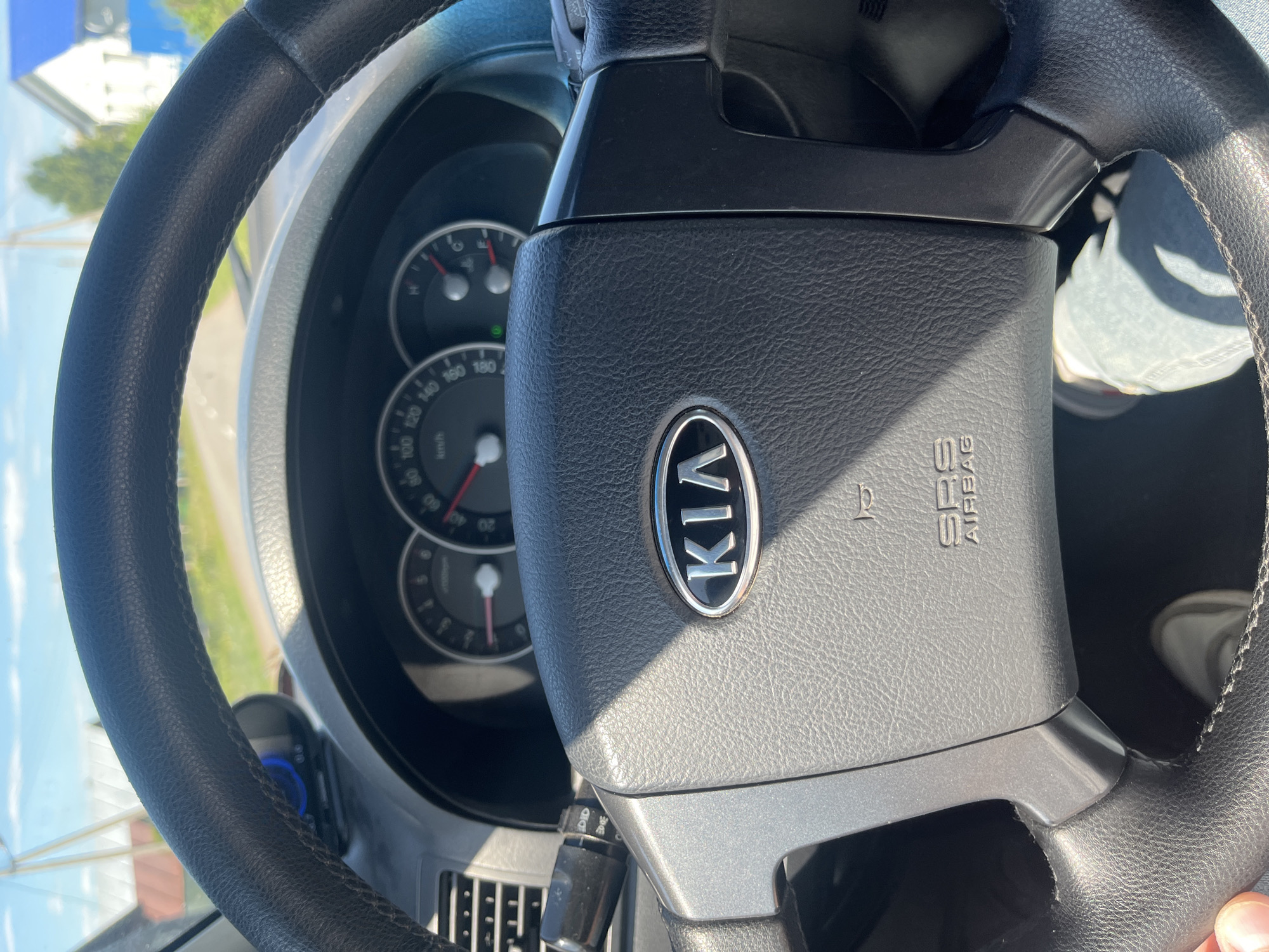 The image shows the interior of a Kia vehicle, viewed from the driver's perspective. The steering wheel, with the Kia logo prominently displayed, is in the foreground. The dashboard features a speedometer and tachometer, with a bright sunny sky visible through the windshield. Outside the car, a rural setting with houses and trees can be seen, suggesting the car is driving on a road near a residential area. The overall impression is that of a casual drive on a clear day in a suburban or rural environment.